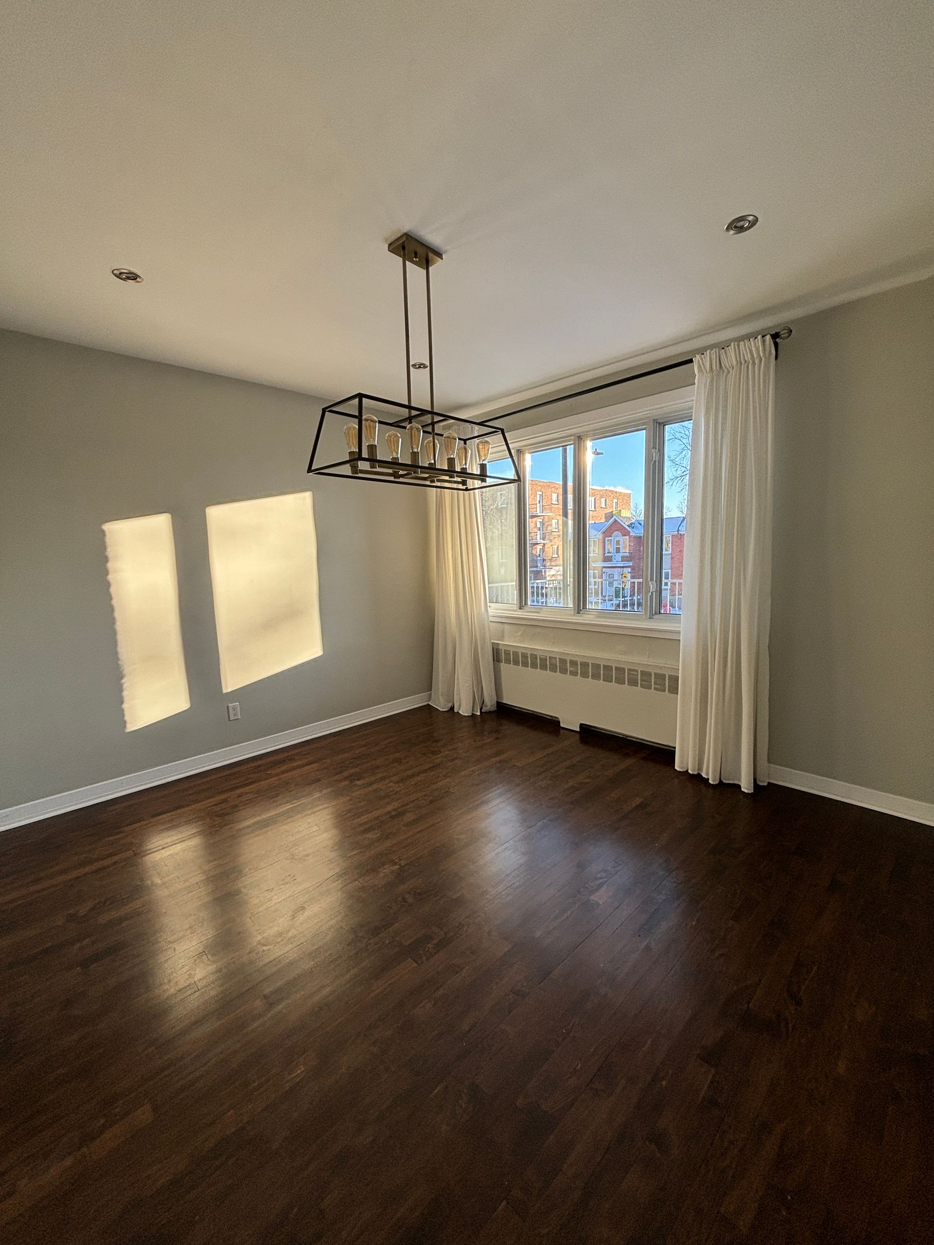 Chambre vide avec parquet, une grande fenêtre avec rideaux blancs et un lustre moderne.