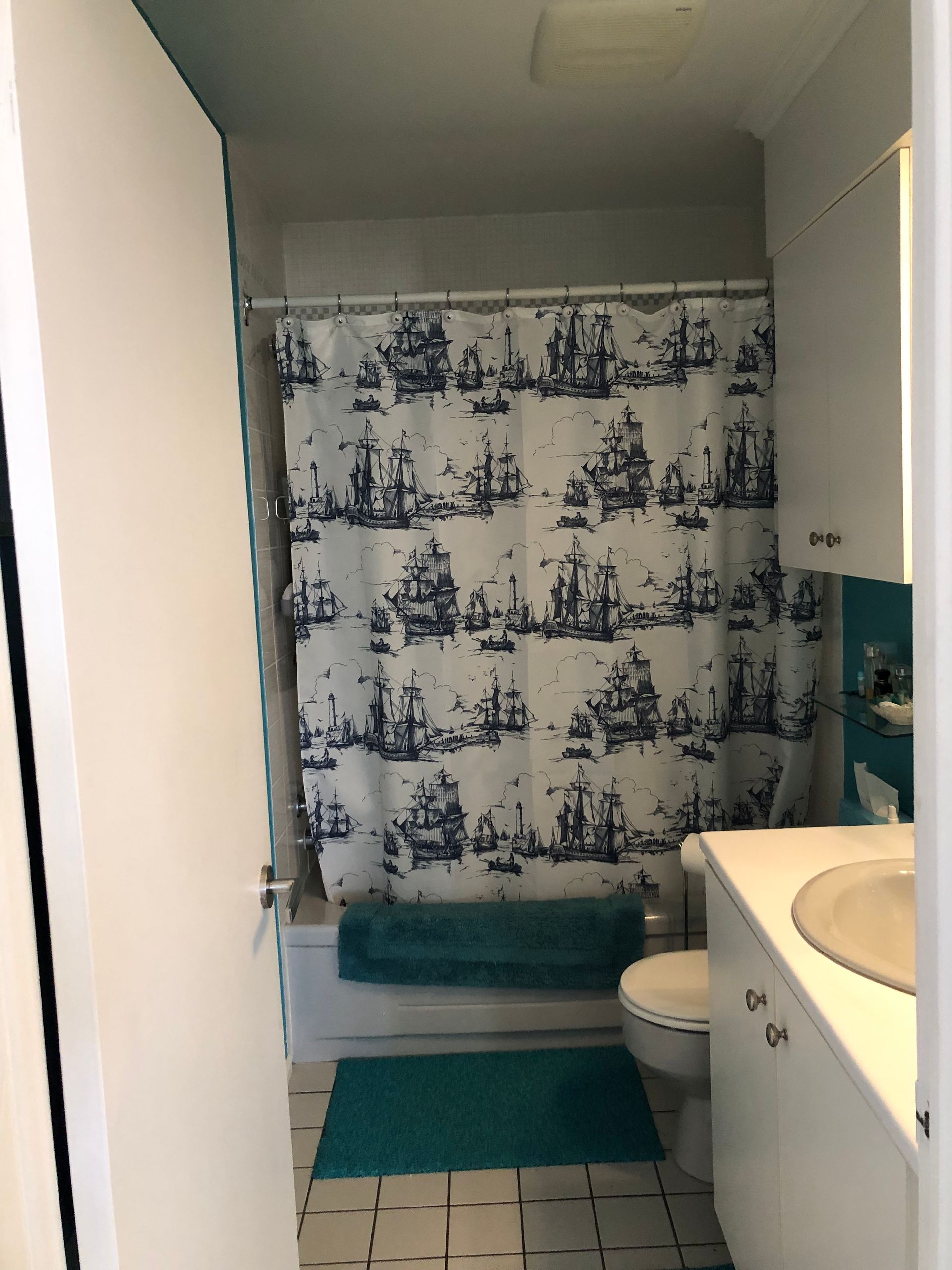 Salle de bain avec un rideau de douche à thème nautique, un tapis de bain bleu sarcelle et une vanité blanche.