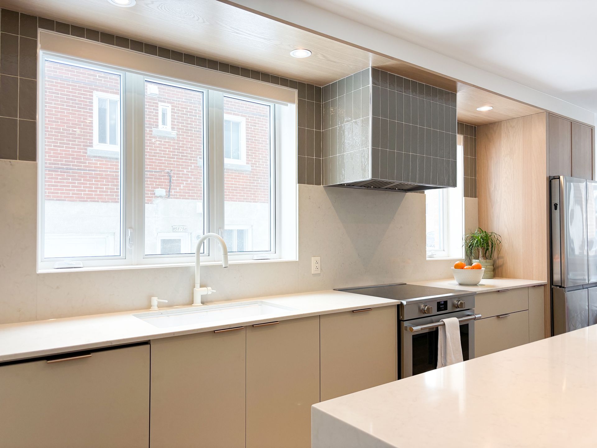 Cuisine moderne avec armoires crème, appareils électroménagers en acier inoxydable et une fenêtre donnant sur un bâtiment en briques.