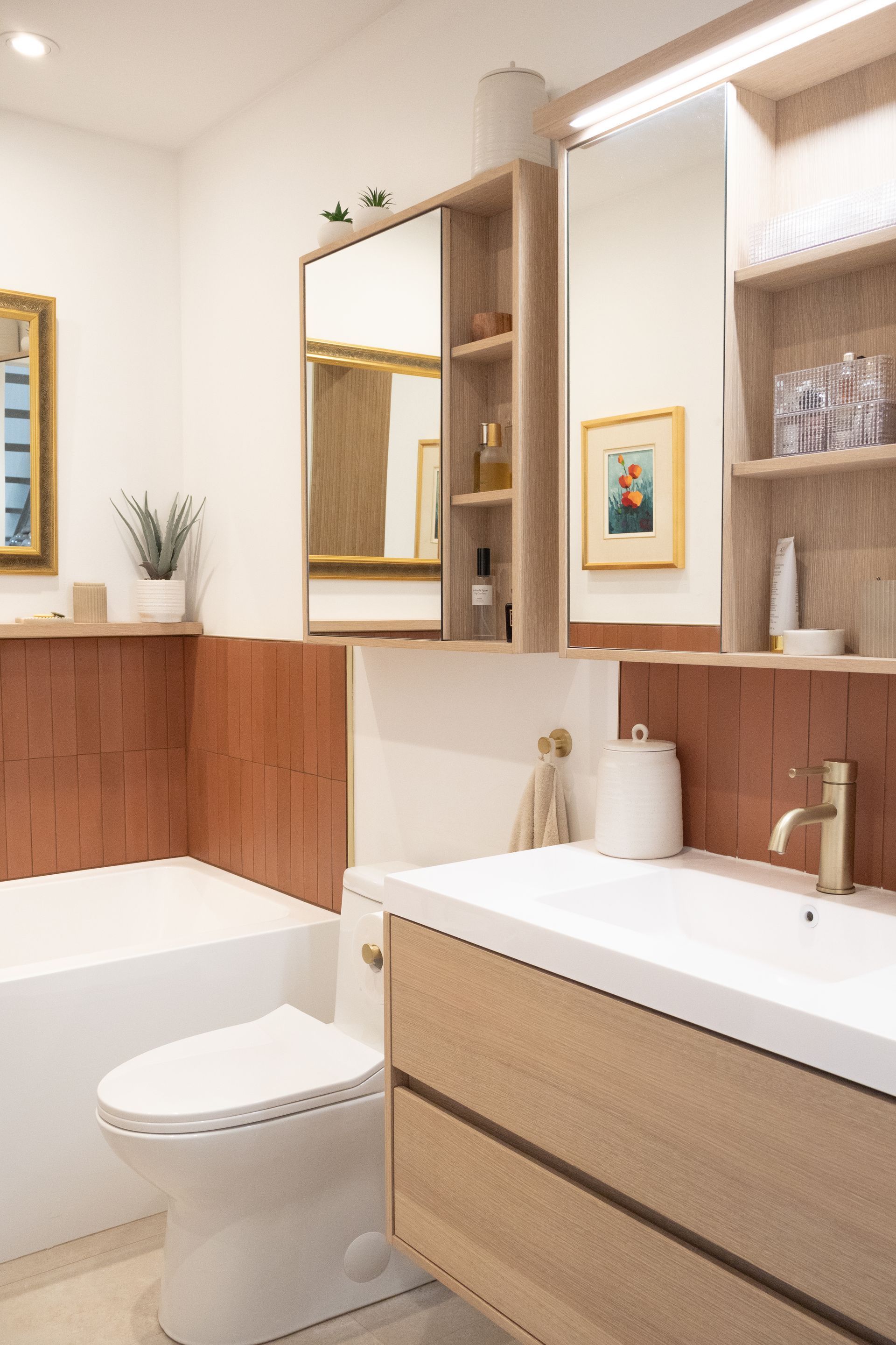 Salle de bain avec murs carrelés couleur terre cuite, lavabo blanc, robinet doré, rangement en bois et œuvres d'art encadrées.