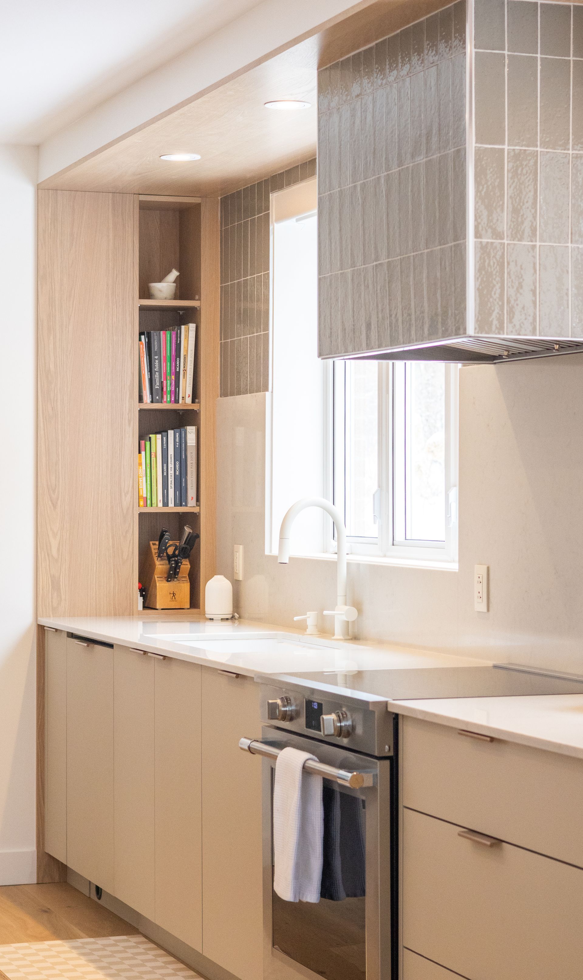Cuisine moderne avec armoires en bois clair, four en acier inoxydable et fenêtre avec dosseret en carrelage gris.