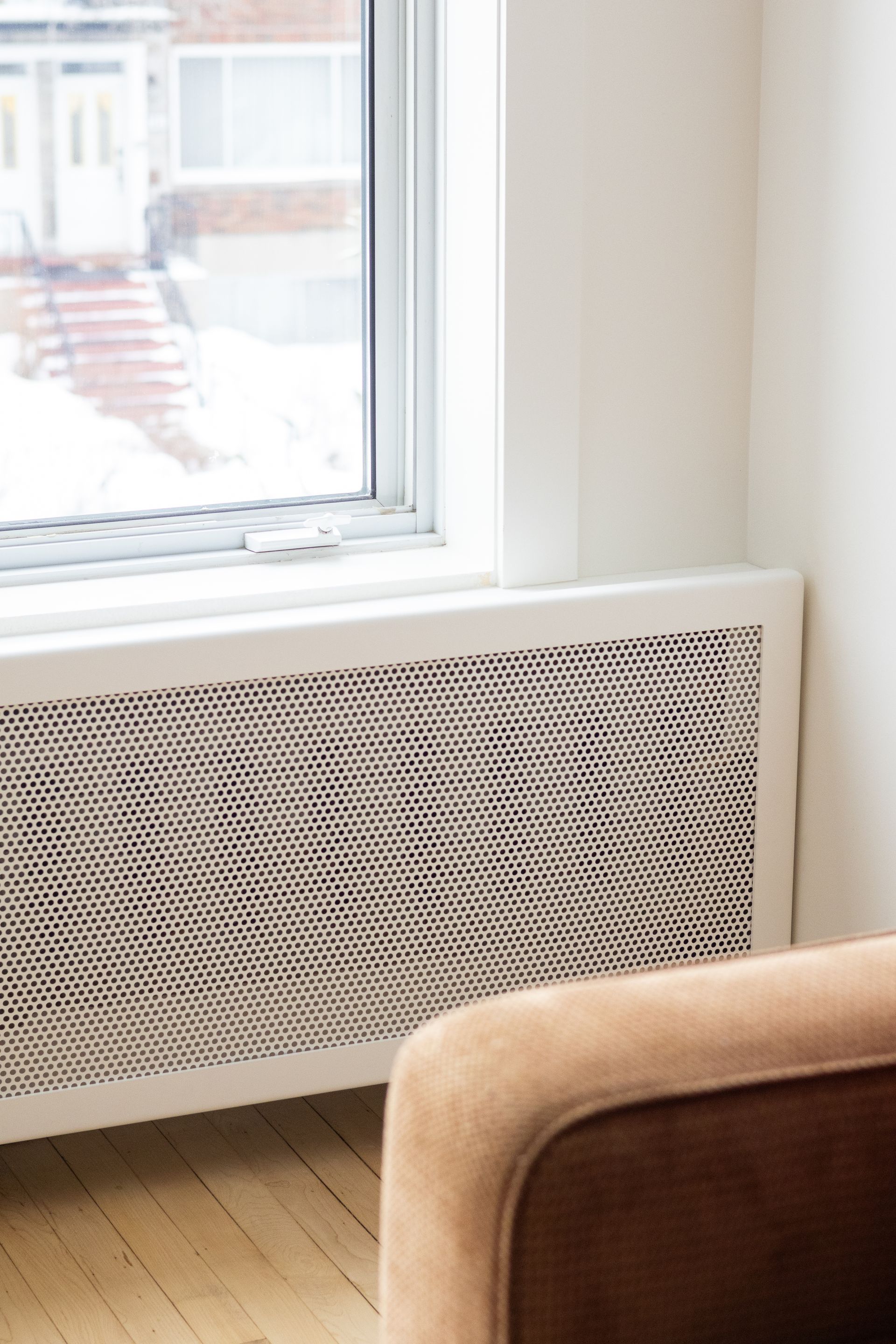 Fenêtre à cadre blanc avec cache-radiateur ; plancher en bois, chaise beige visible.