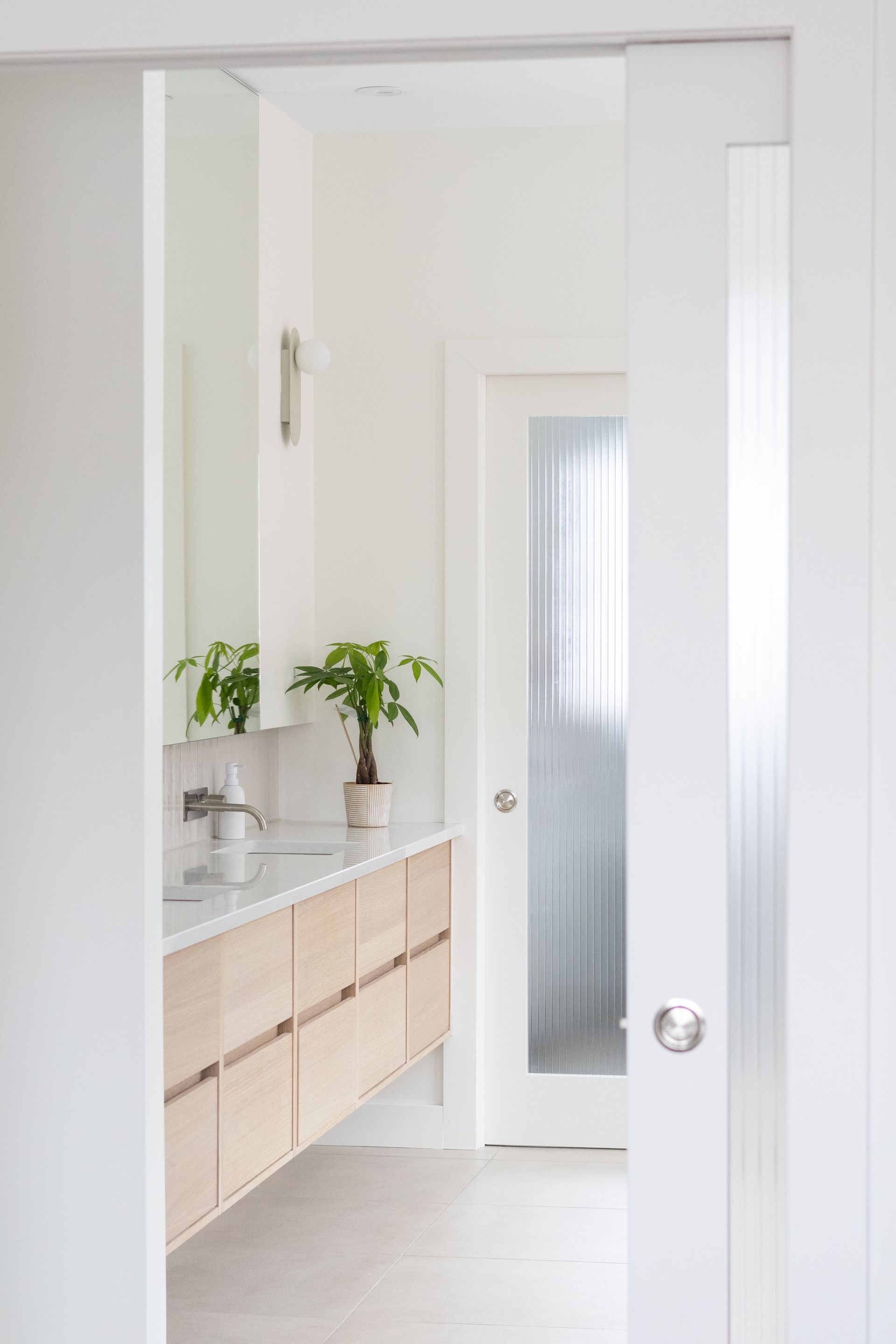 Intérieur de la salle de bain : meuble-lavabo en bois clair, murs blancs, porte en verre dépoli, cadre de porte ouvert.