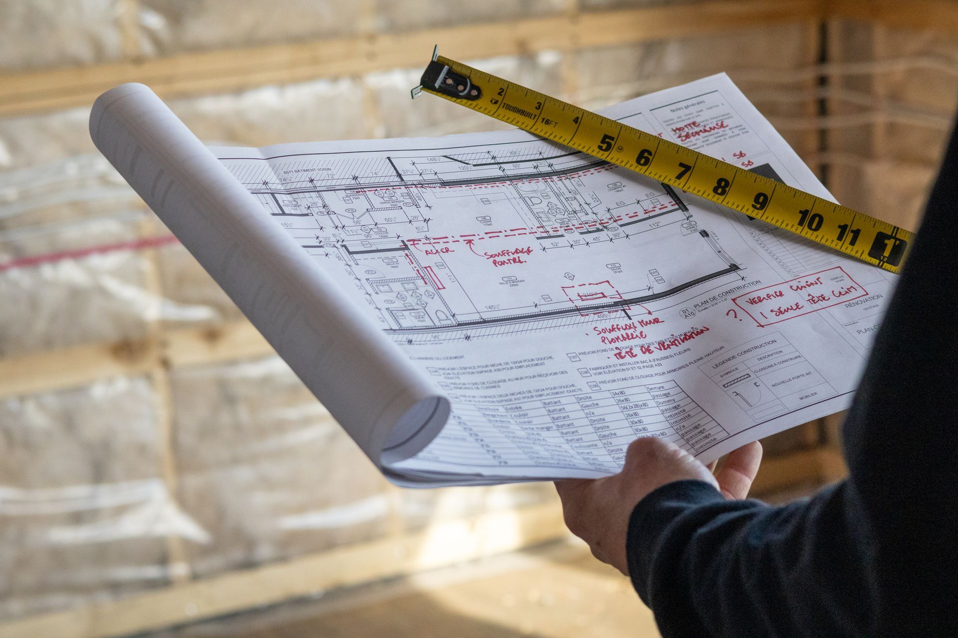 Personne tenant des plans enroulés avec un ruban à mesurer dessus, à l'intérieur d'un bâtiment en construction.