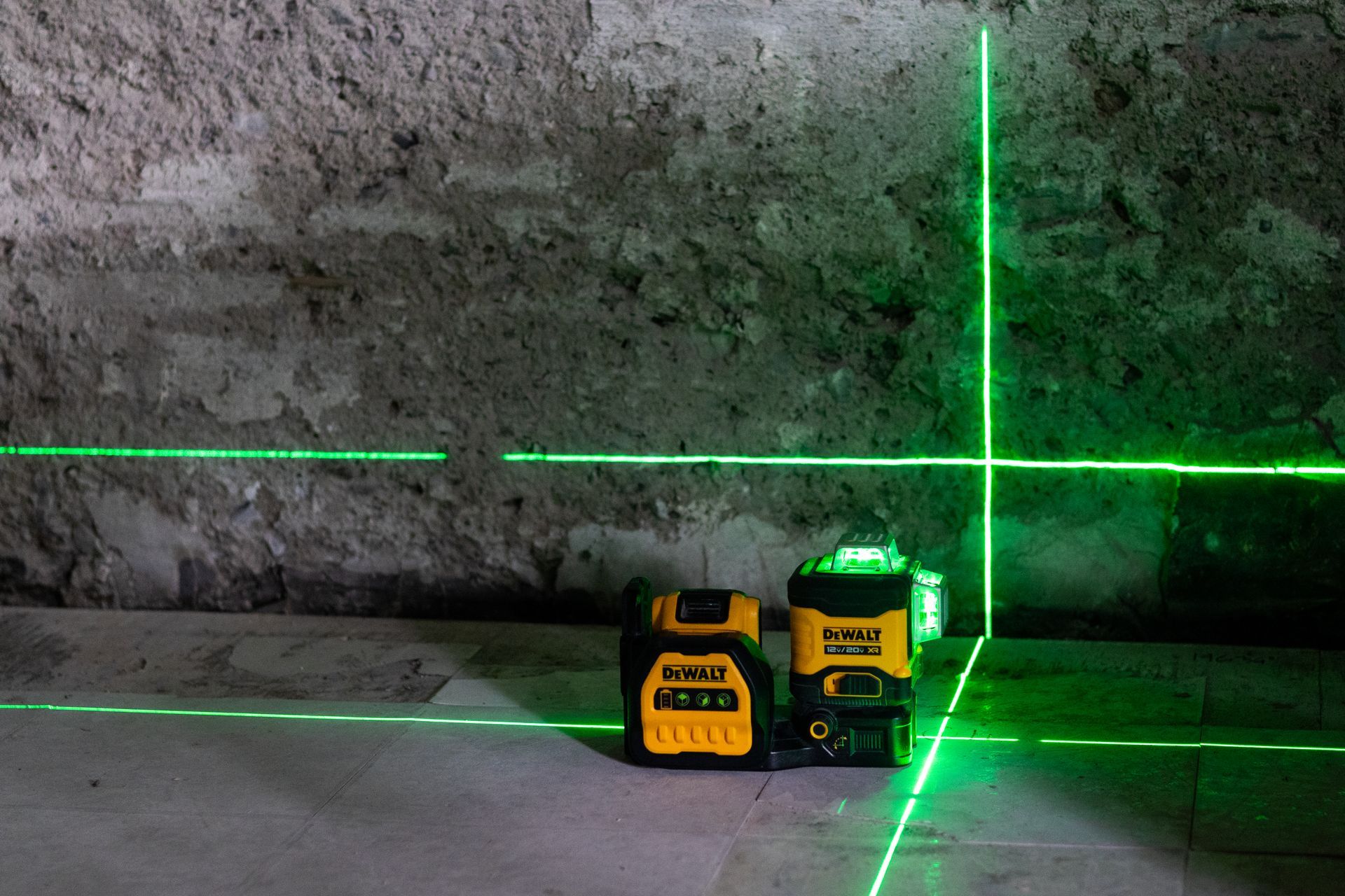 Deux niveaux laser verts projetant des lignes qui se croisent sur un mur en béton brut.