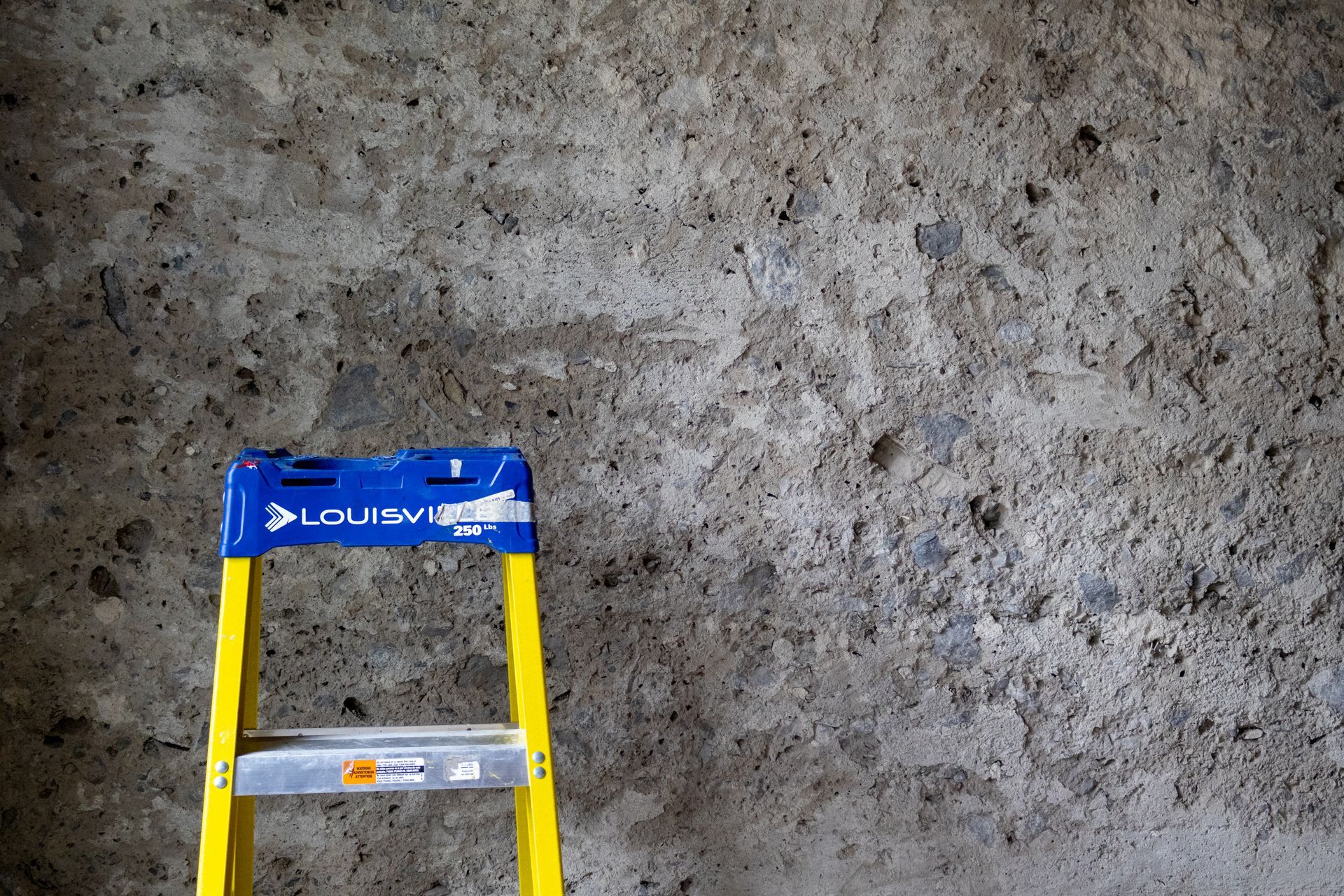 Escabeau jaune et bleu appuyé contre un mur de béton gris et rugueux.