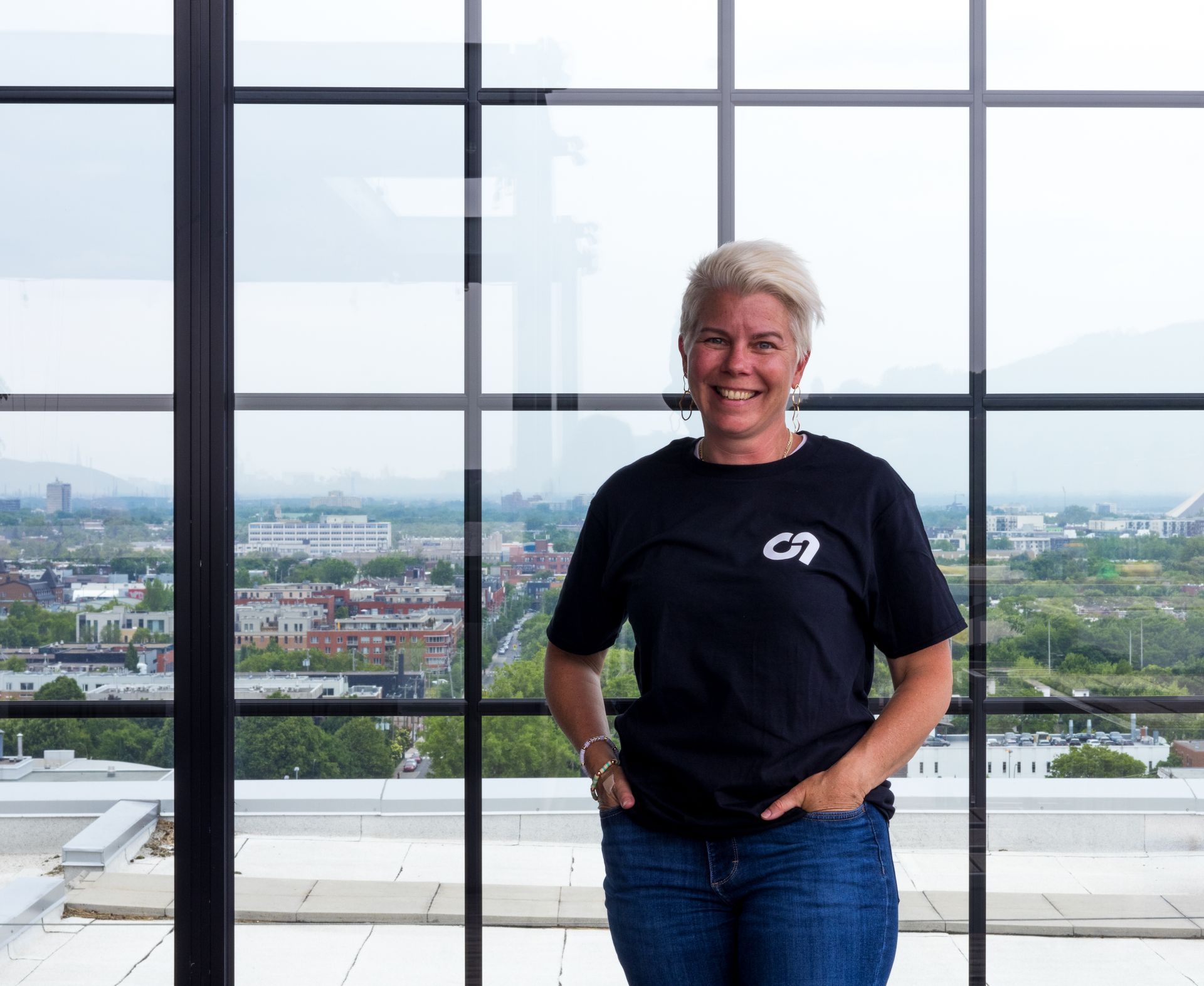 Une femme aux cheveux blonds courts en t-shirt noir et jean se tient devant une fenêtre donnant sur une ville.