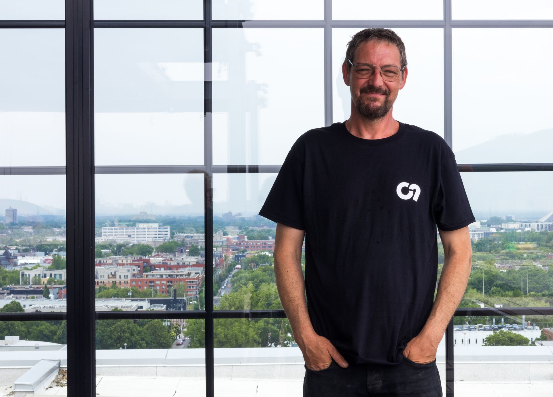 Un homme en t-shirt noir, les mains dans les poches, se tient près d'une fenêtre donnant sur un paysage urbain.