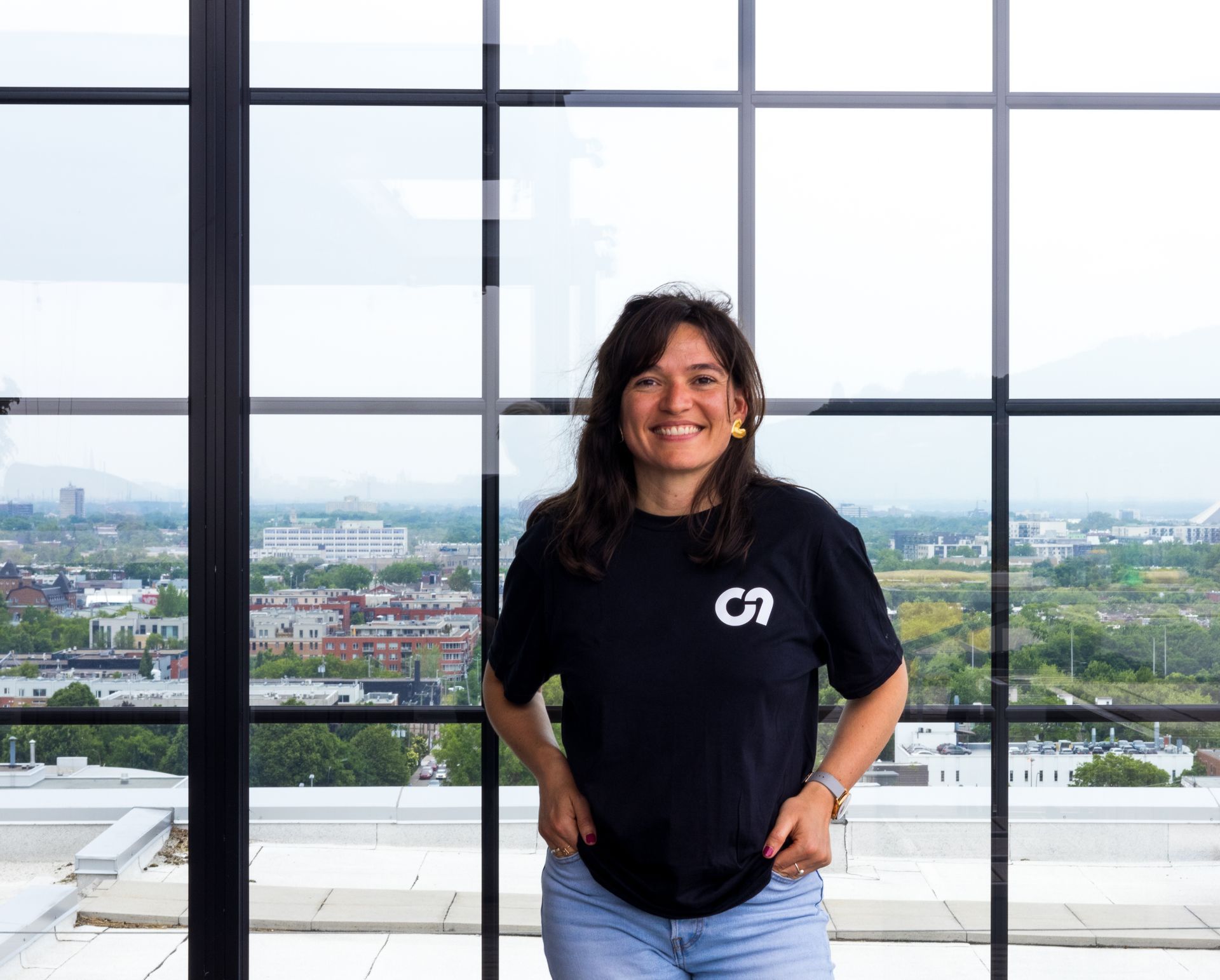 Femme portant une chemise noire avec un logo, souriant devant une fenêtre avec vue sur la ville.