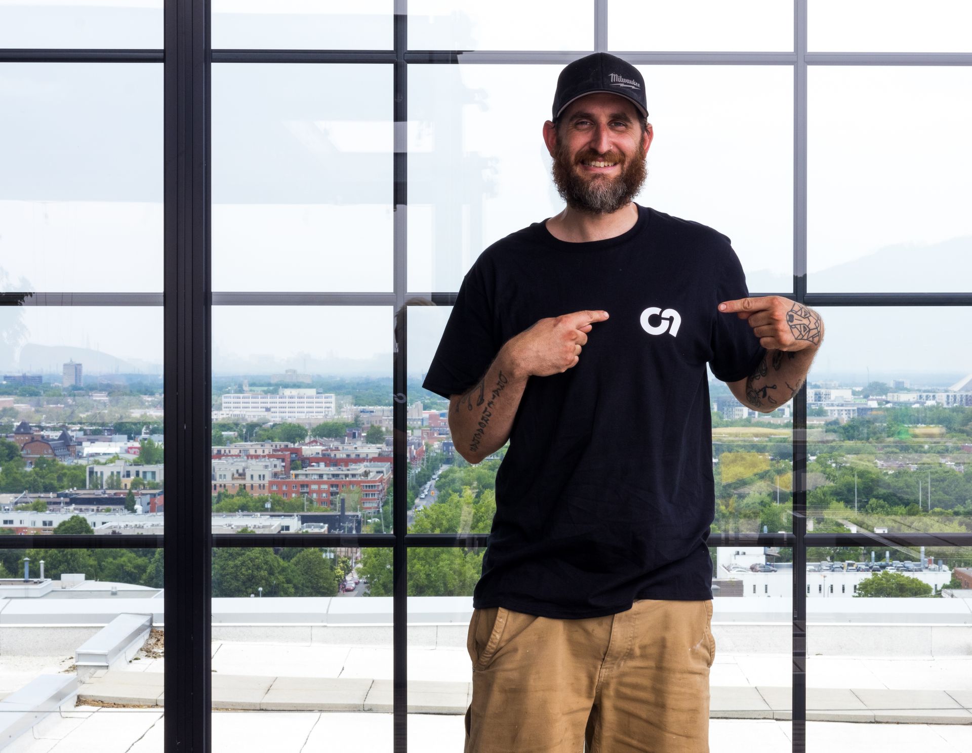 Un homme en chemise noire avec le logo « CN » pointe son t-shirt du doigt. Il porte une casquette et se tient près d'une fenêtre avec vue sur la ville.