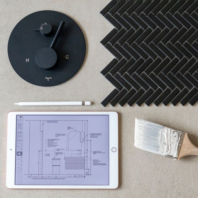 Robinet de douche noir, carrelage à chevrons, plan numérique sur tablette, pinceau et crayon sur une surface grise.