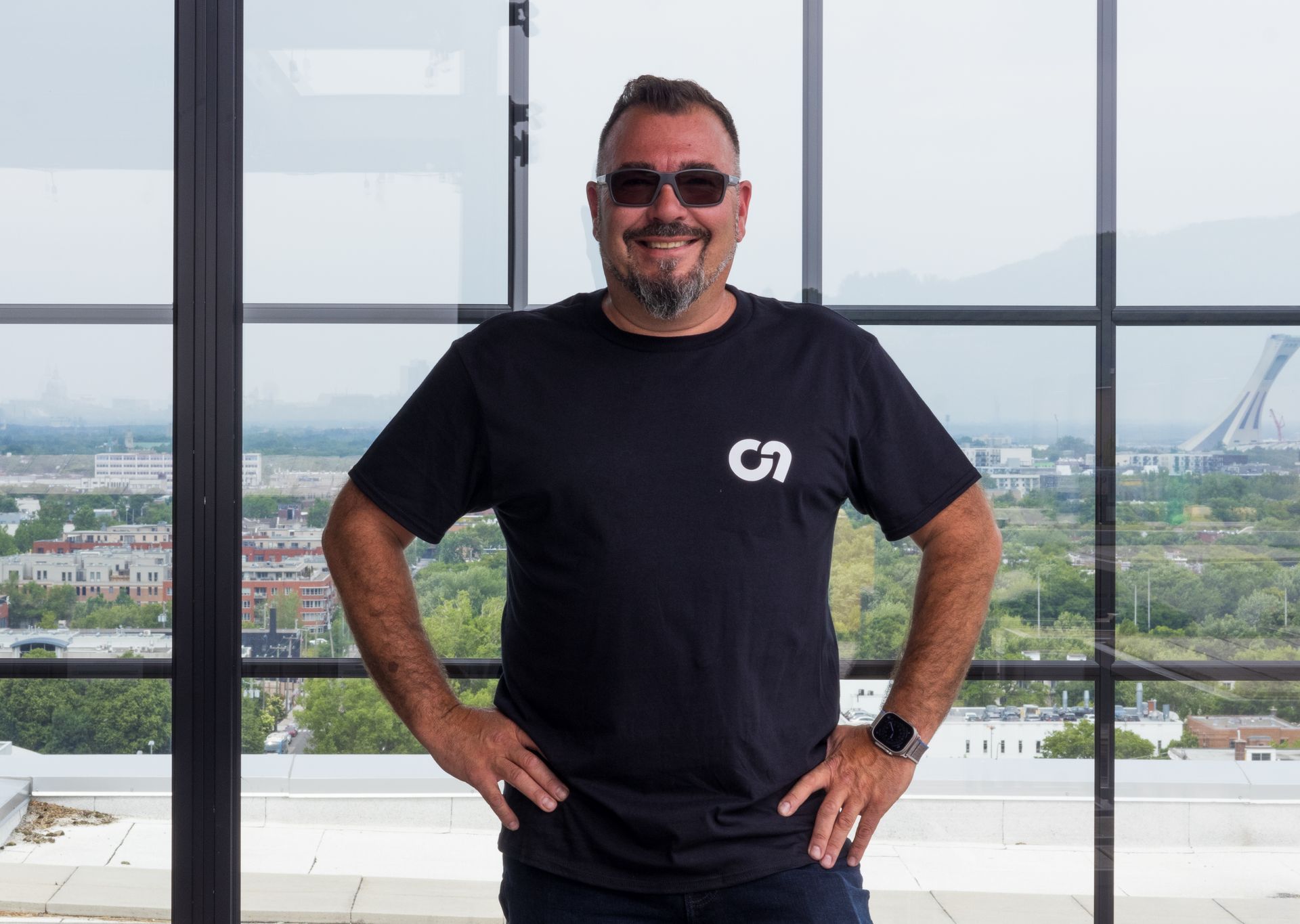 Homme en chemise noire et lunettes de soleil, les mains sur les hanches, souriant, à l'extérieur près de la vue sur la ville.