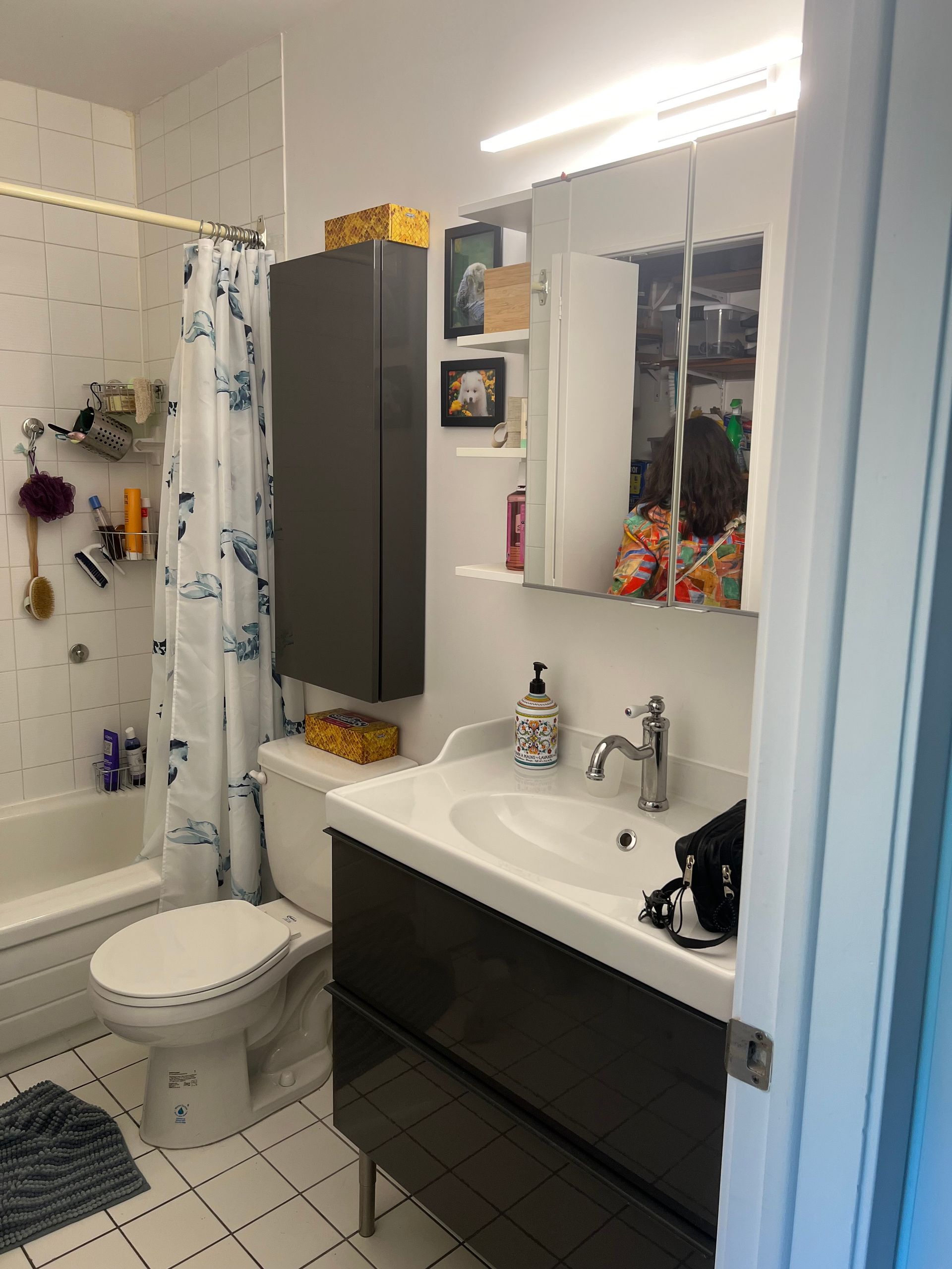 Une petite salle de bain avec toilettes, lavabo, douche et meuble de rangement. Tons blancs et gris.