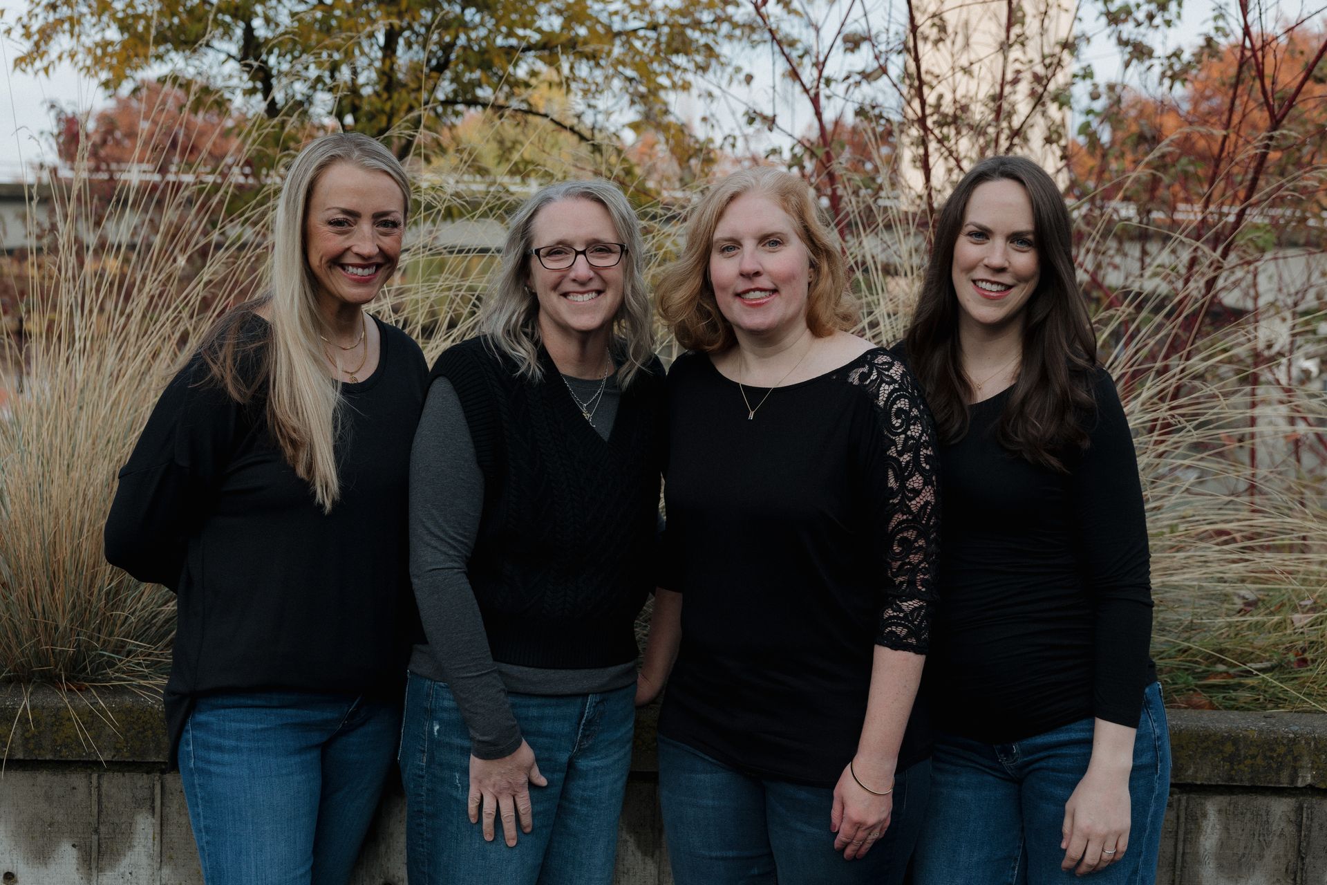 Four women smiling outdoors; one wearing glasses, others with blonde or red hair; dark tops.