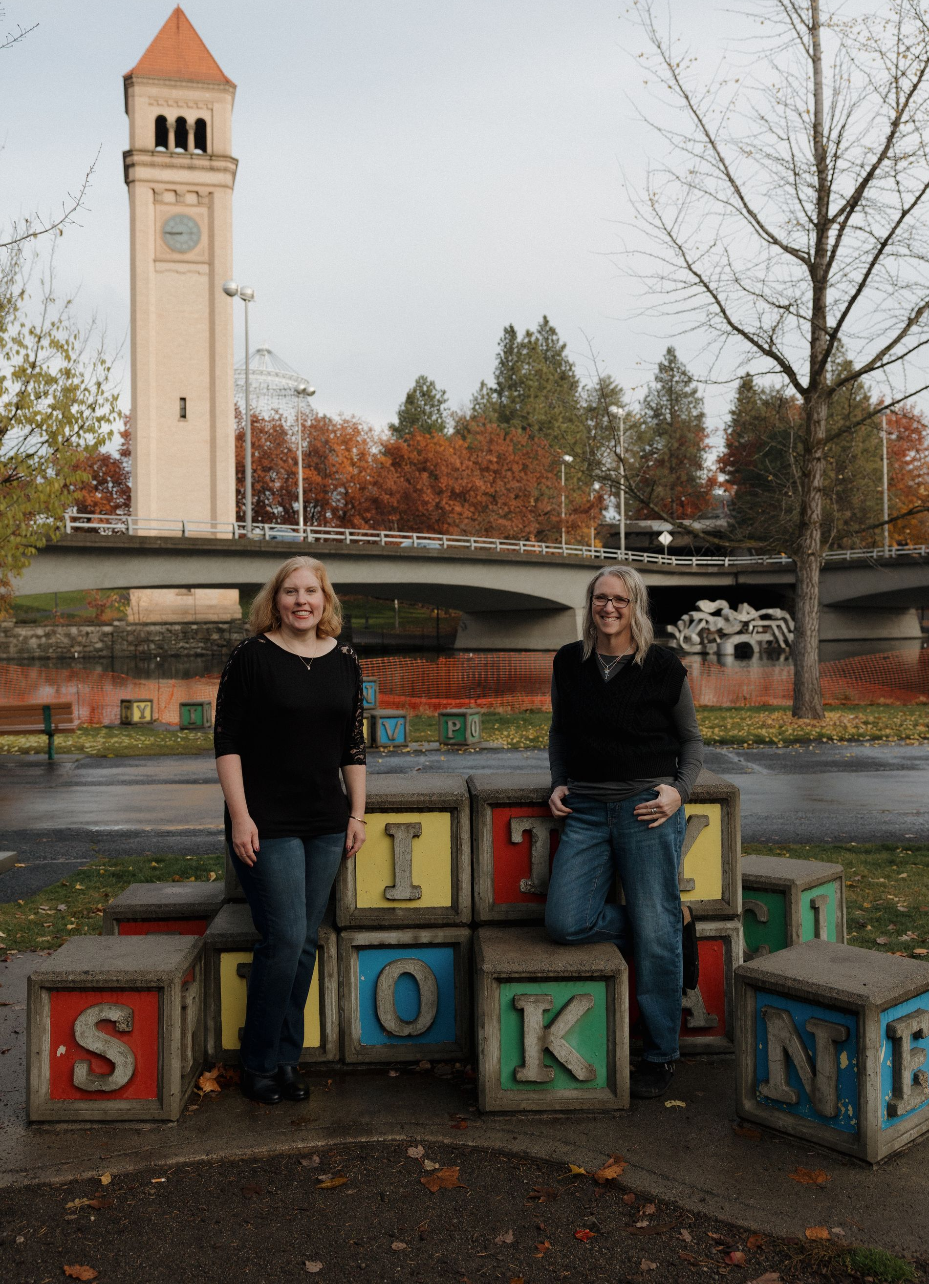 Two women in matching teal sweaters smile, arms crossed, standing back-to-back outdoors with fall foliage.