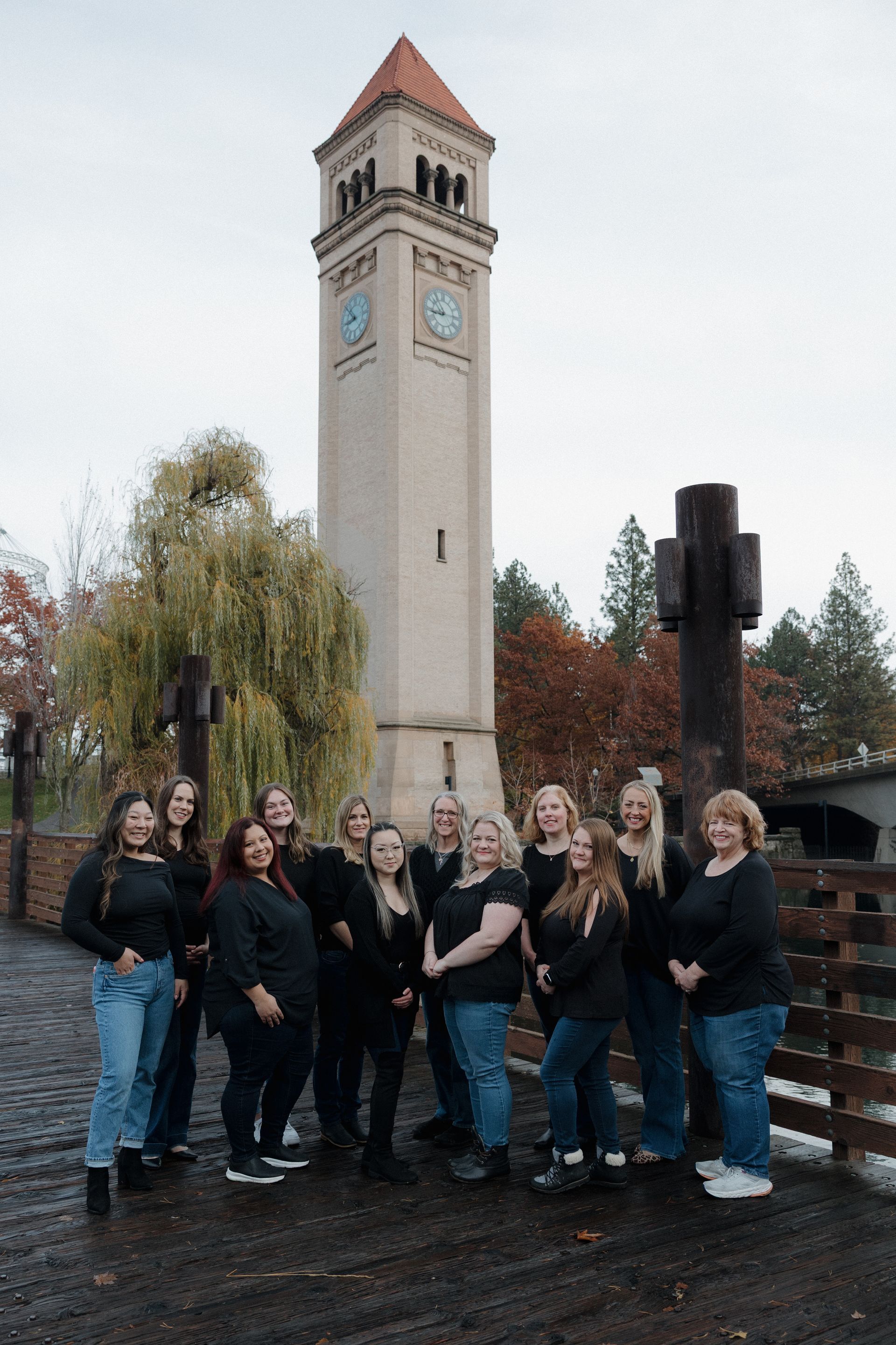 Four women smiling, posing outdoors. Green foliage background, varying hair colors and tops.