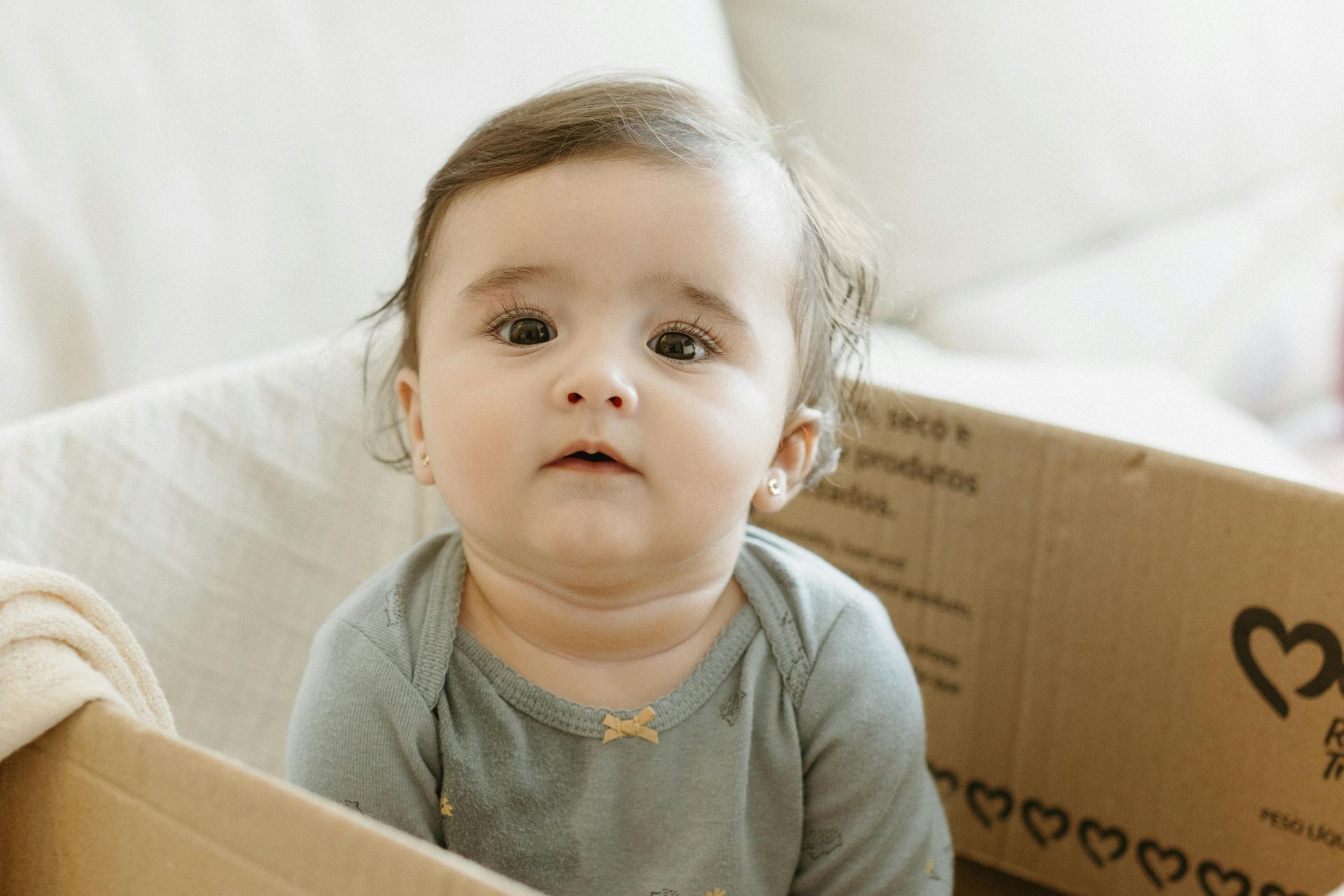 A baby girl is sitting in a cardboard box looking at the camera.