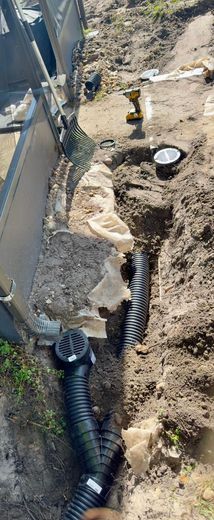 Trench with black corrugated drain pipes, a drainage catch basin, and a drill, likely for drainage installation.