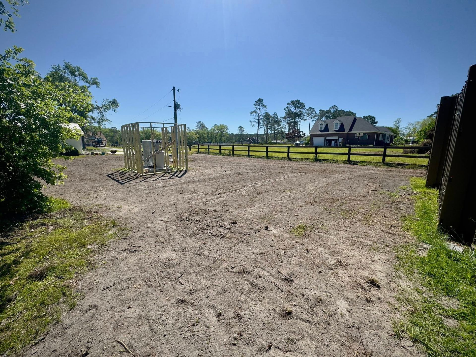 Cleared dirt lot with a wooden structure, fence, and house under a bright sky.