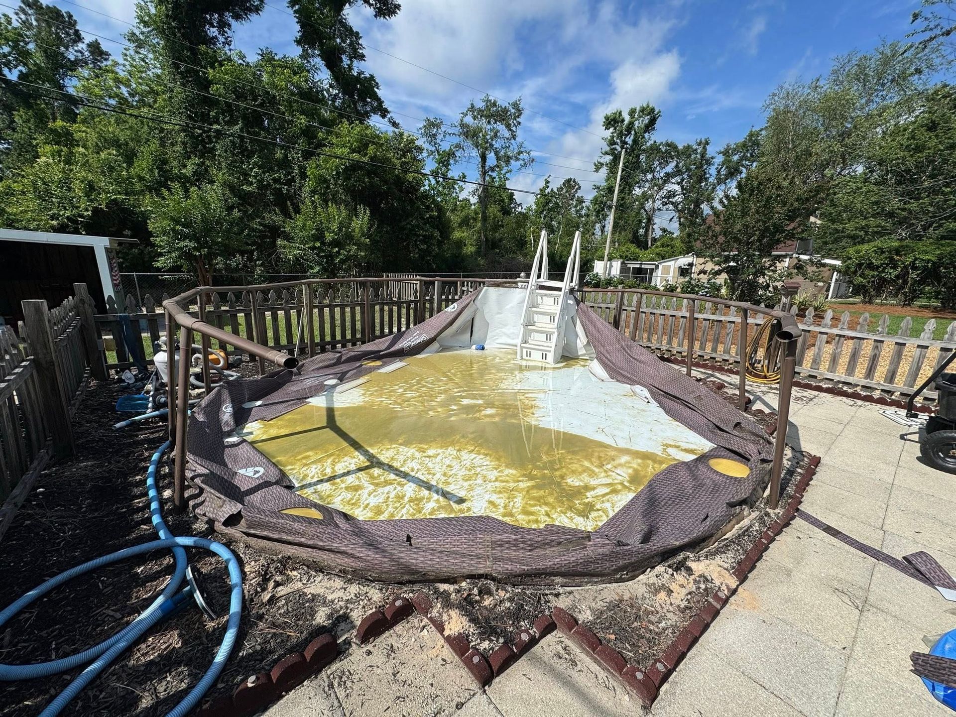 Dilapidated above-ground pool with murky water and damaged liner, set in a backyard with a wooden fence.