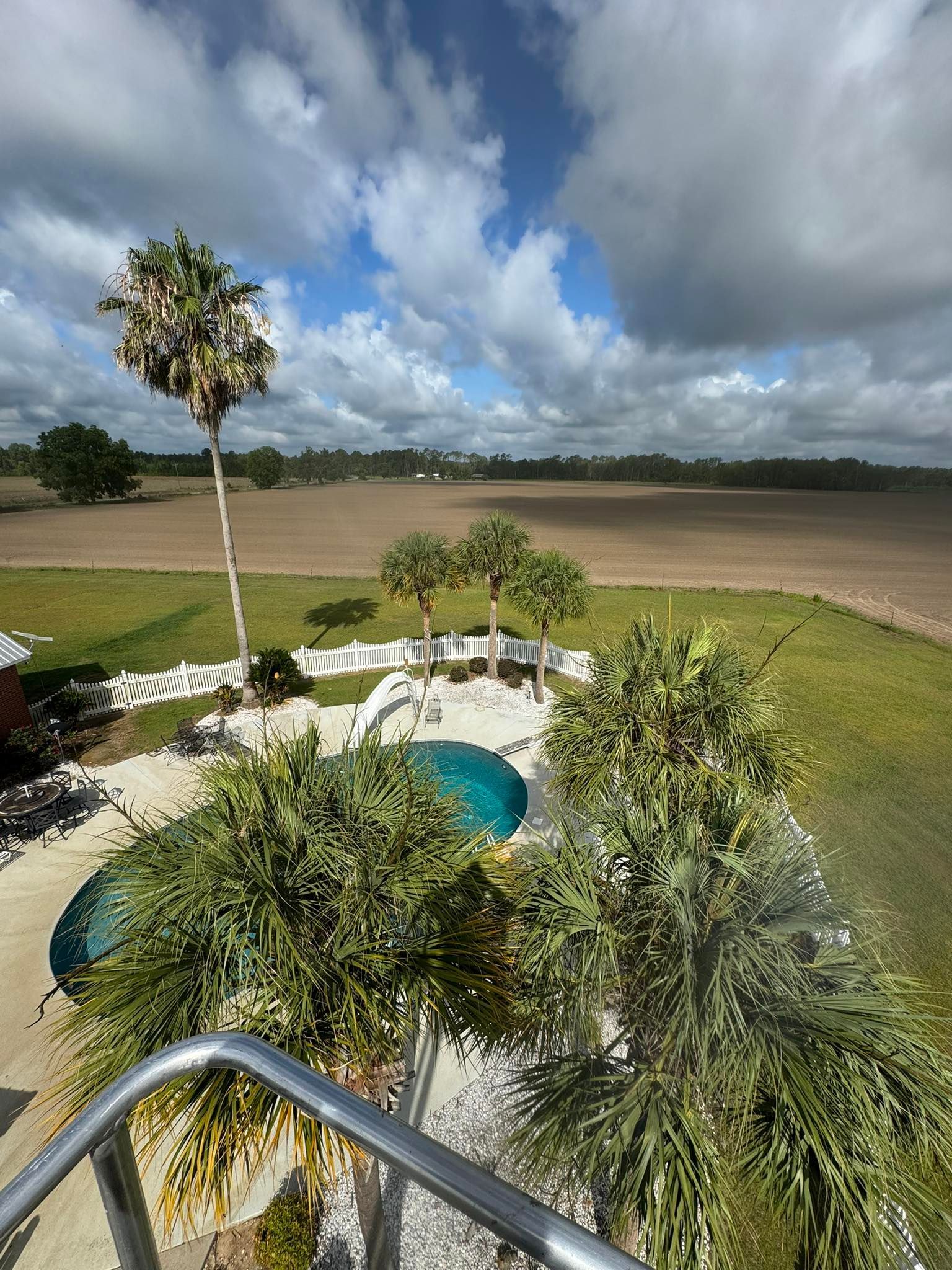 Overlooking a pool and lawn with a palm tree, and a farm field under a cloudy sky.