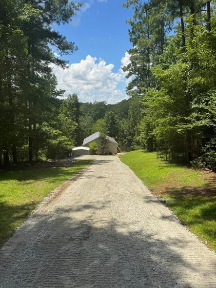 Gravel driveway leading to a building in a wooded area under a blue sky with clouds.