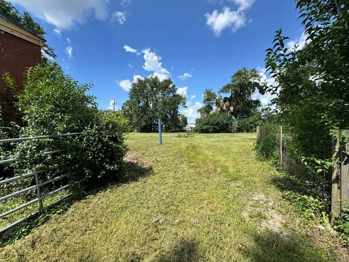 Grassy lot with blue markers under a partly cloudy sky, flanked by trees and a metal fence.