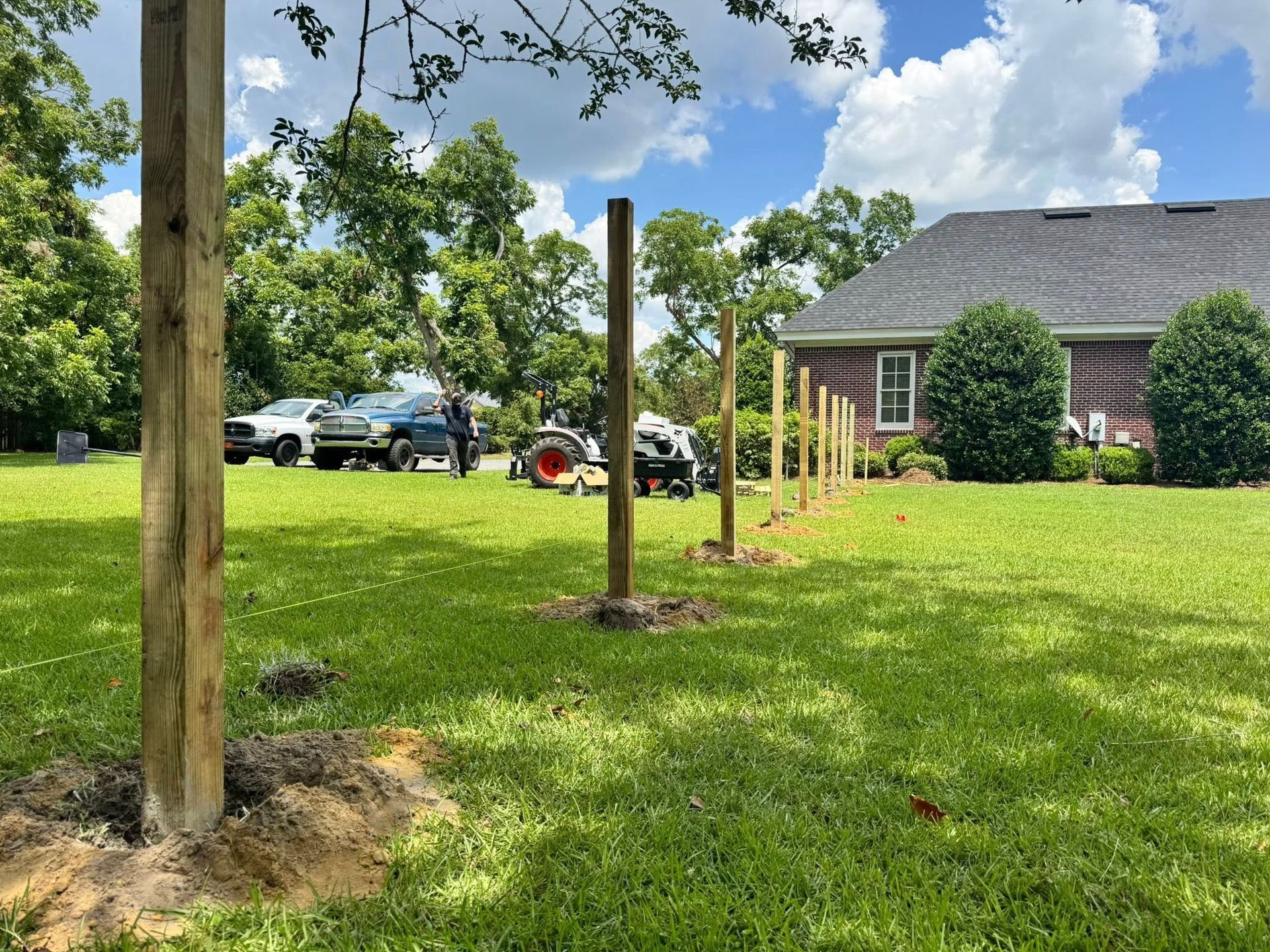 Building a fence in a grassy yard. Wooden posts are erected in a row, with a tractor and vehicles nearby.
