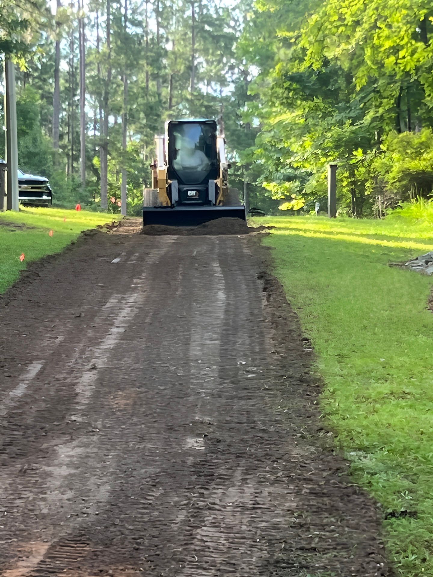A skid steer loader levels dirt on a residential driveway between grassy lawns and trees.