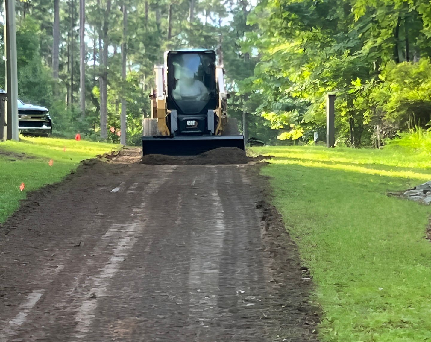 Skid steer grading a dirt road with green grass on both sides, trees in the background.