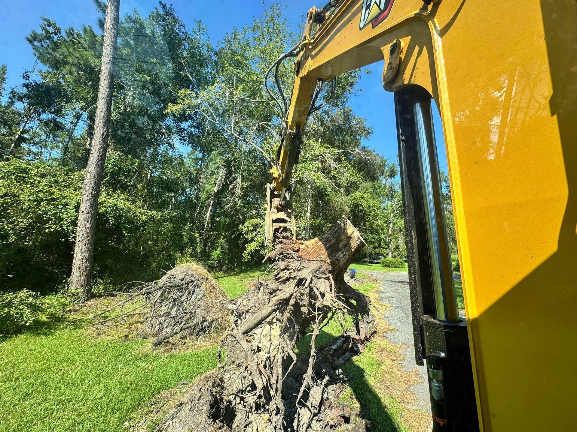 Yellow excavator digging in a grassy area, removing dirt and roots near trees and a path.
