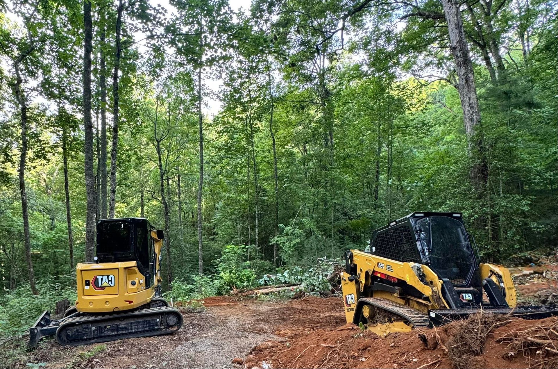 Two construction vehicles in a wooded area; a yellow excavator and a yellow skid steer.