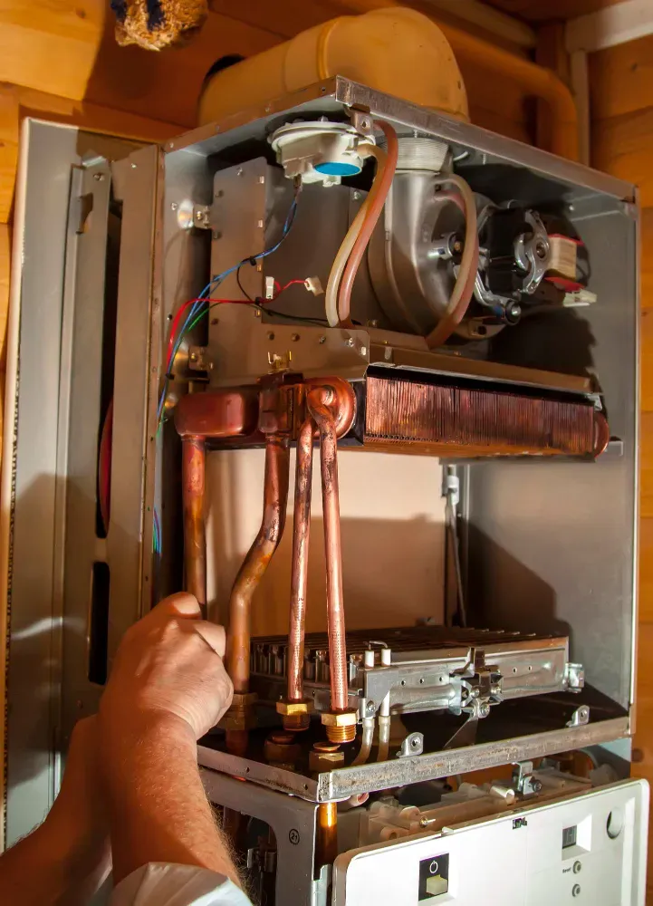Hands working on a gas boiler, copper pipes visible inside the metal casing.