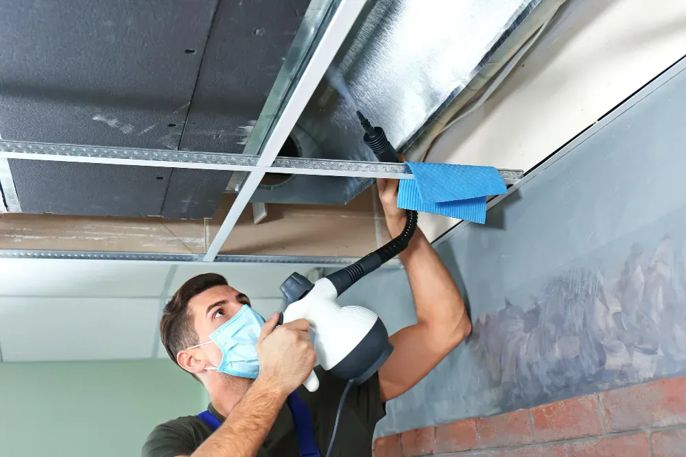 Person in mask uses a steamer to clean ceiling tiles and metal framing.