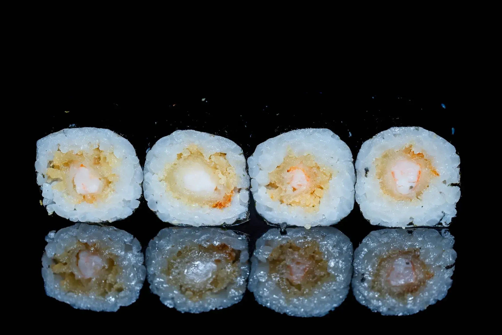 Quattro rotoli di sushi, disposti su due file su sfondo nero, con la fila inferiore che appare più scura e riflettente.