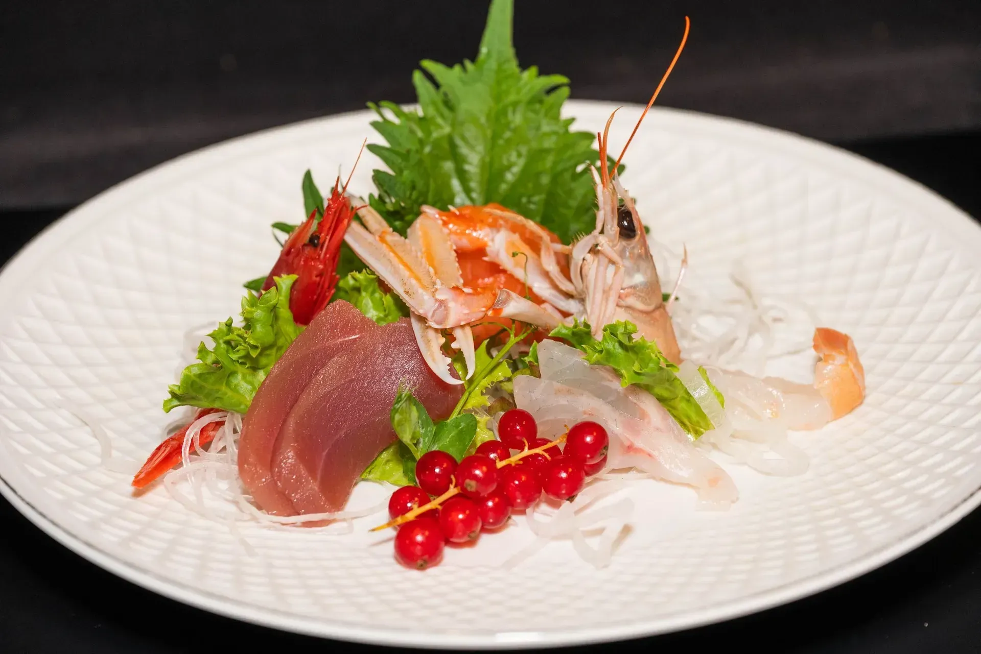 Piatto di sashimi di pesce con tonno, gamberi, verdure e frutti di bosco rossi su un piatto bianco.