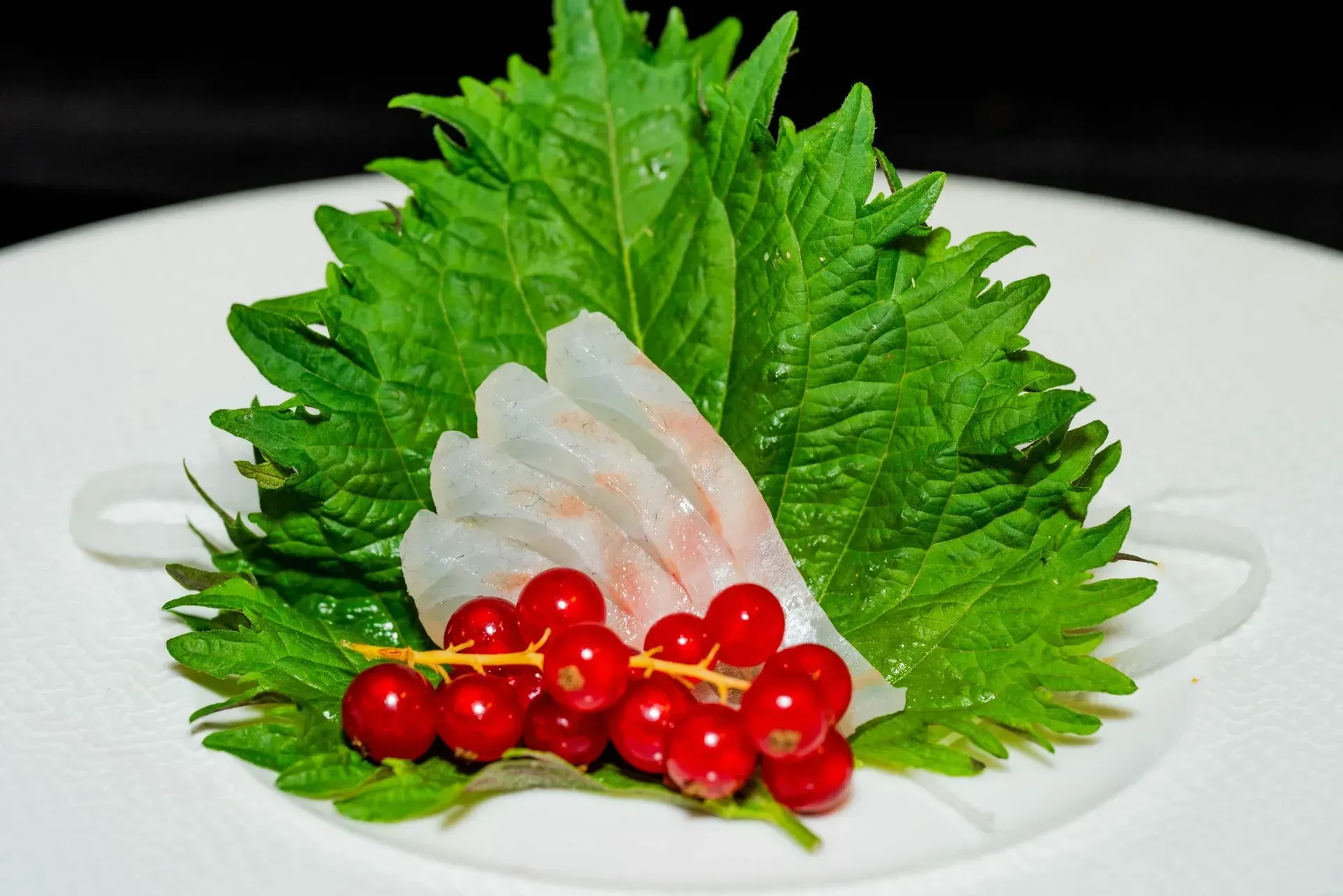 Sashimi guarnito con foglie verdi di shiso, fette di pesce bianco e frutti di bosco rossi su un piatto bianco.