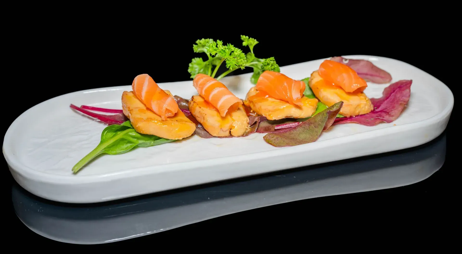 Sushi nigiri disposto su un piatto bianco con guarnizione verde e salsa viola su sfondo nero.