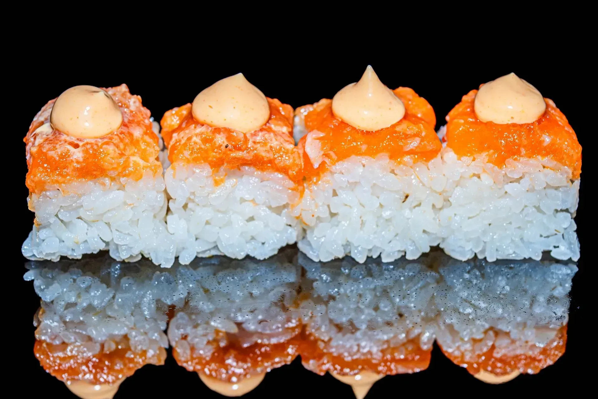 Quattro pezzi di sushi arancioni e bianchi ricoperti di salsa cremosa su una superficie nera riflettente