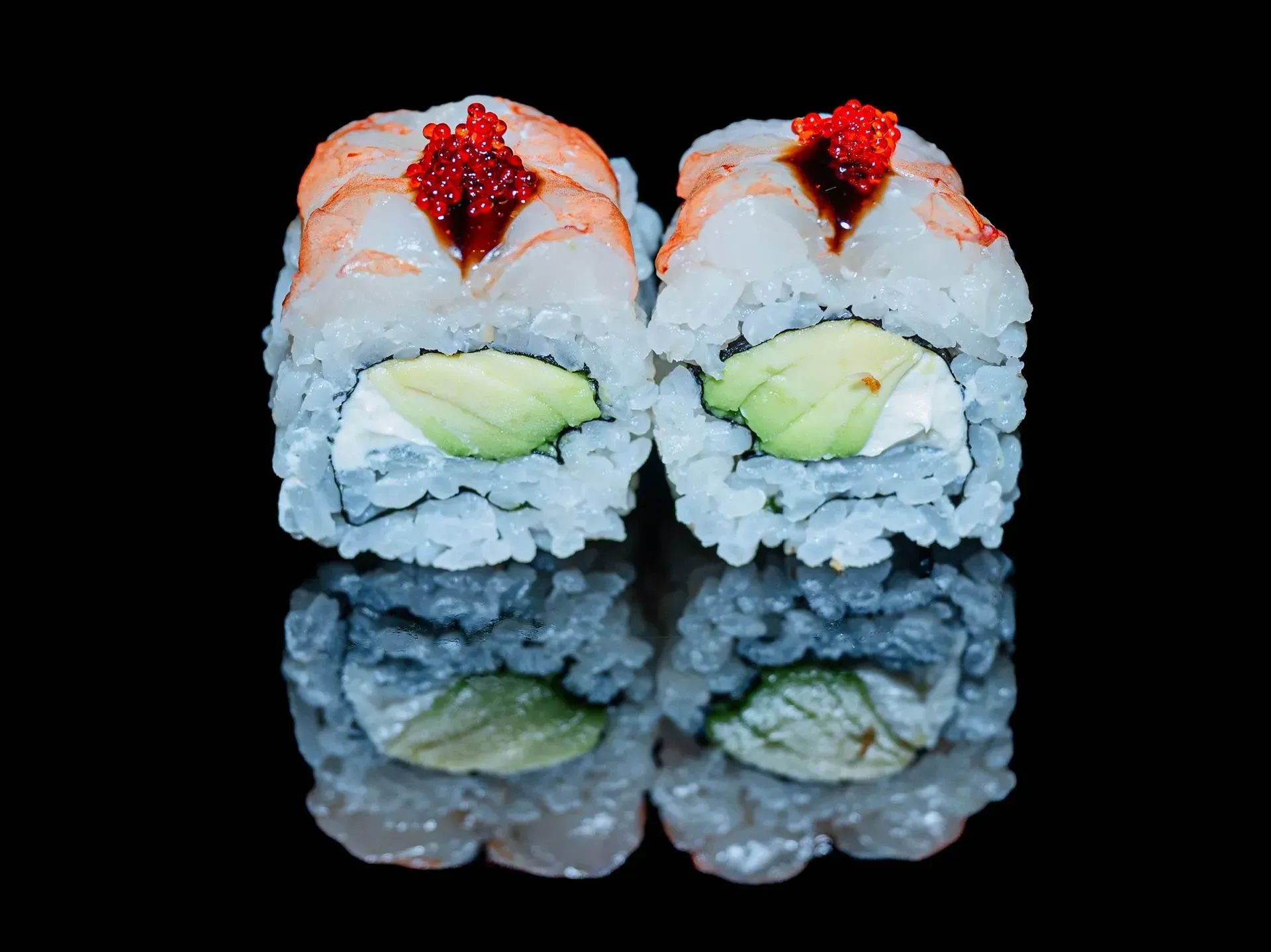 Due rotoli di sushi con avocado e uova di pesce rosse in cima, riflessi su una superficie nera lucida