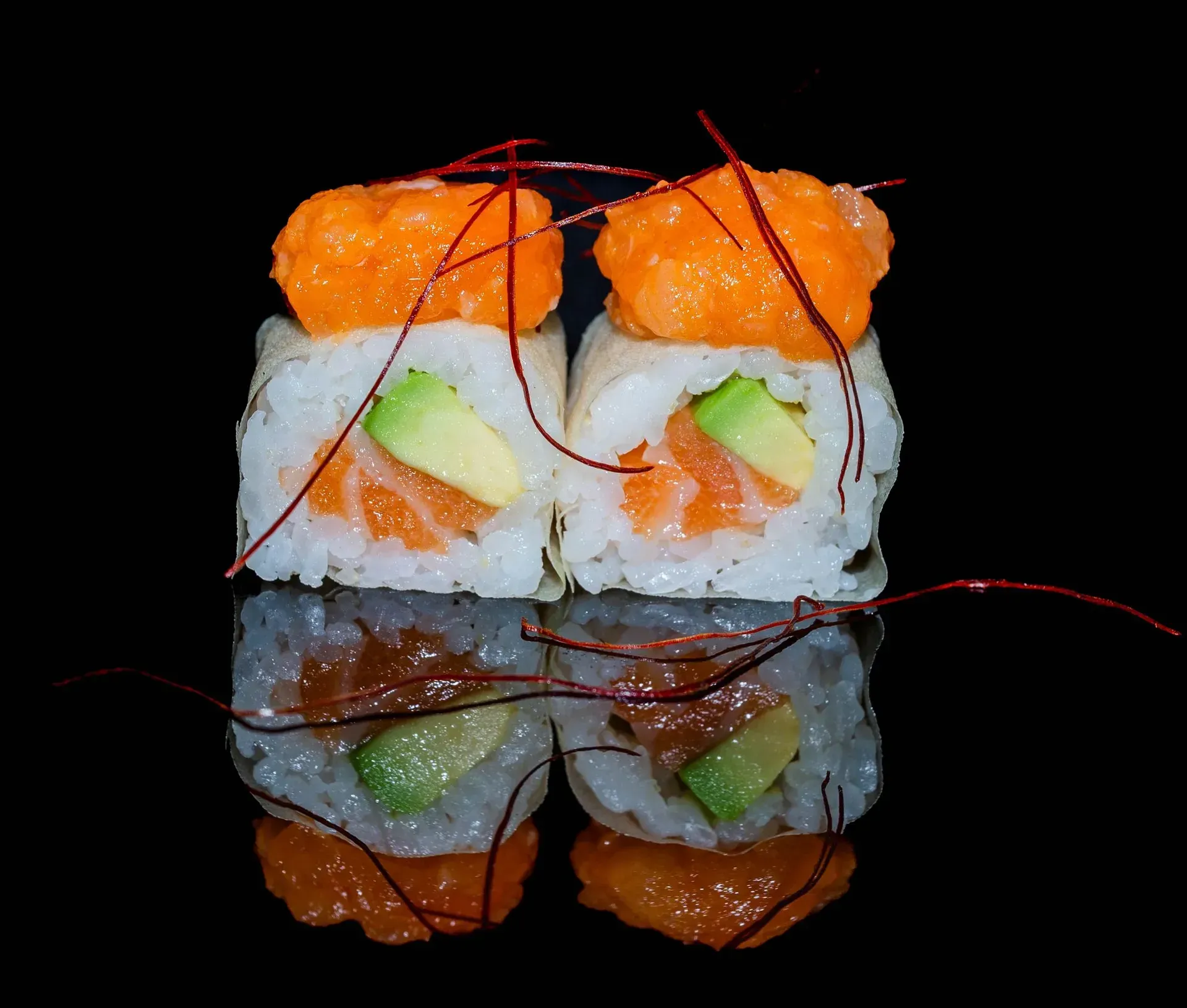 Due rotoli di sushi con guarnizione arancione e ripieno verde, legati con sottili fili rossi su sfondo nero.