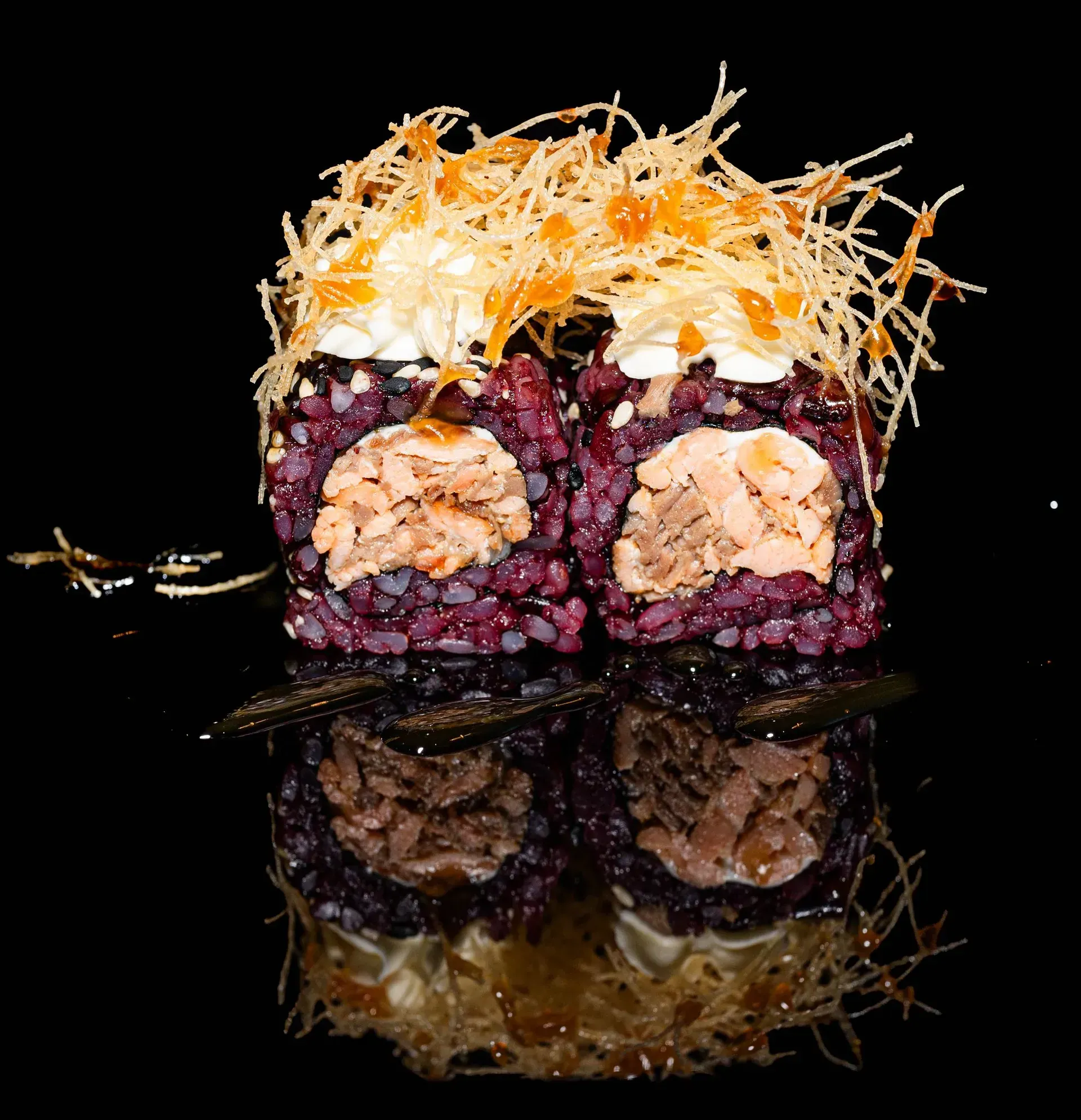 Rotolo di sushi con riso viola, ripieno di salmone e guarnizione di verdure sminuzzate su una superficie nera riflettente.