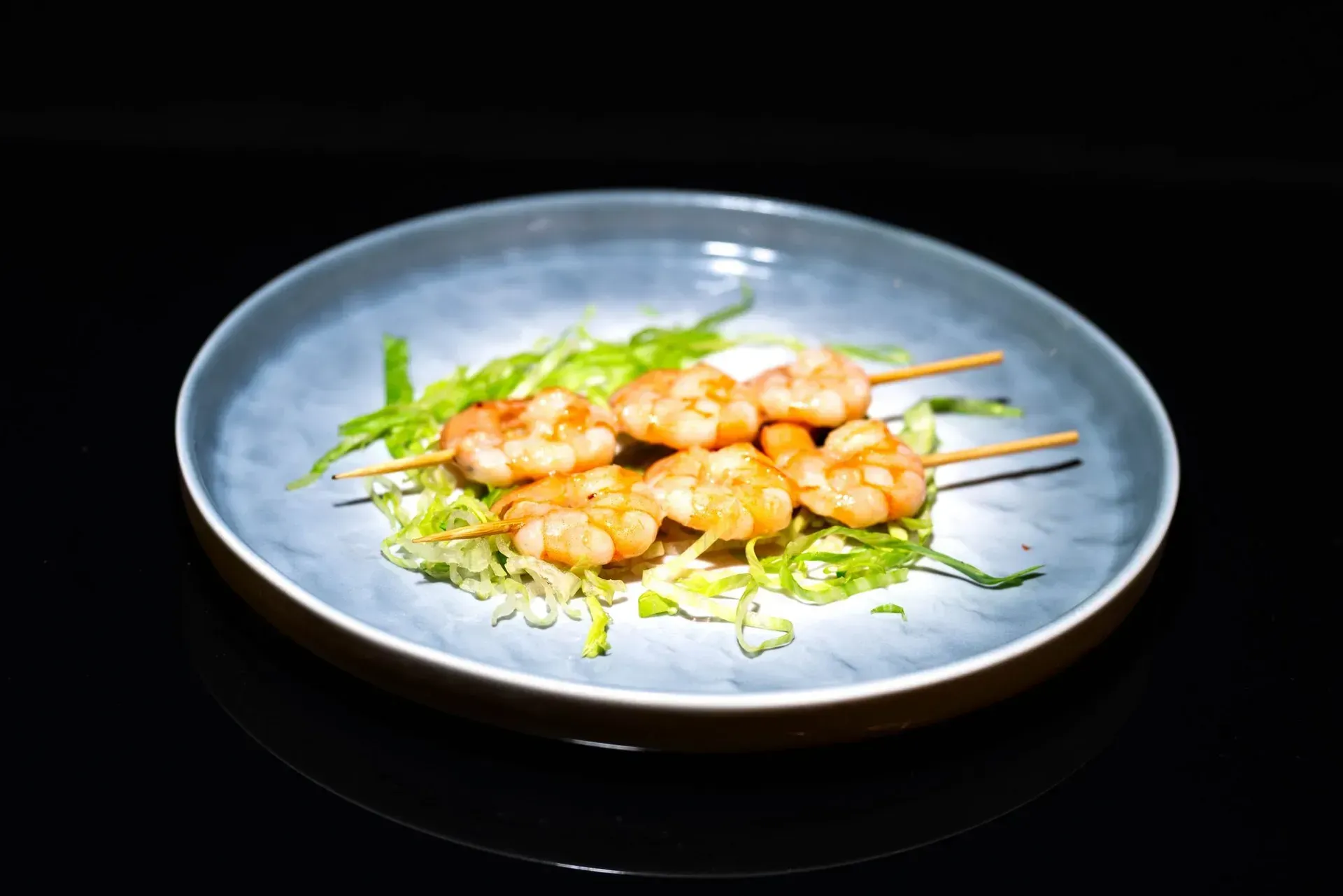 Spiedini di gamberi grigliati su un piatto blu con guarnizione verde su sfondo nero.