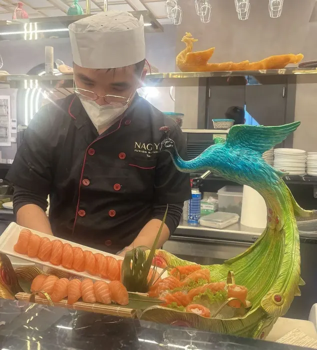 Uno chef con la mascherina prepara il sushi accanto a un piatto colorato a forma di pesce, di colore blu-verde.