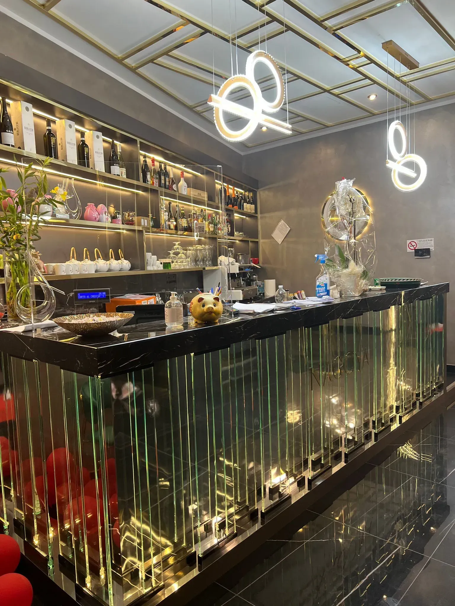 Interni di un bar moderno con scaffali illuminati, un bancone nero e luci a soffitto brillanti.