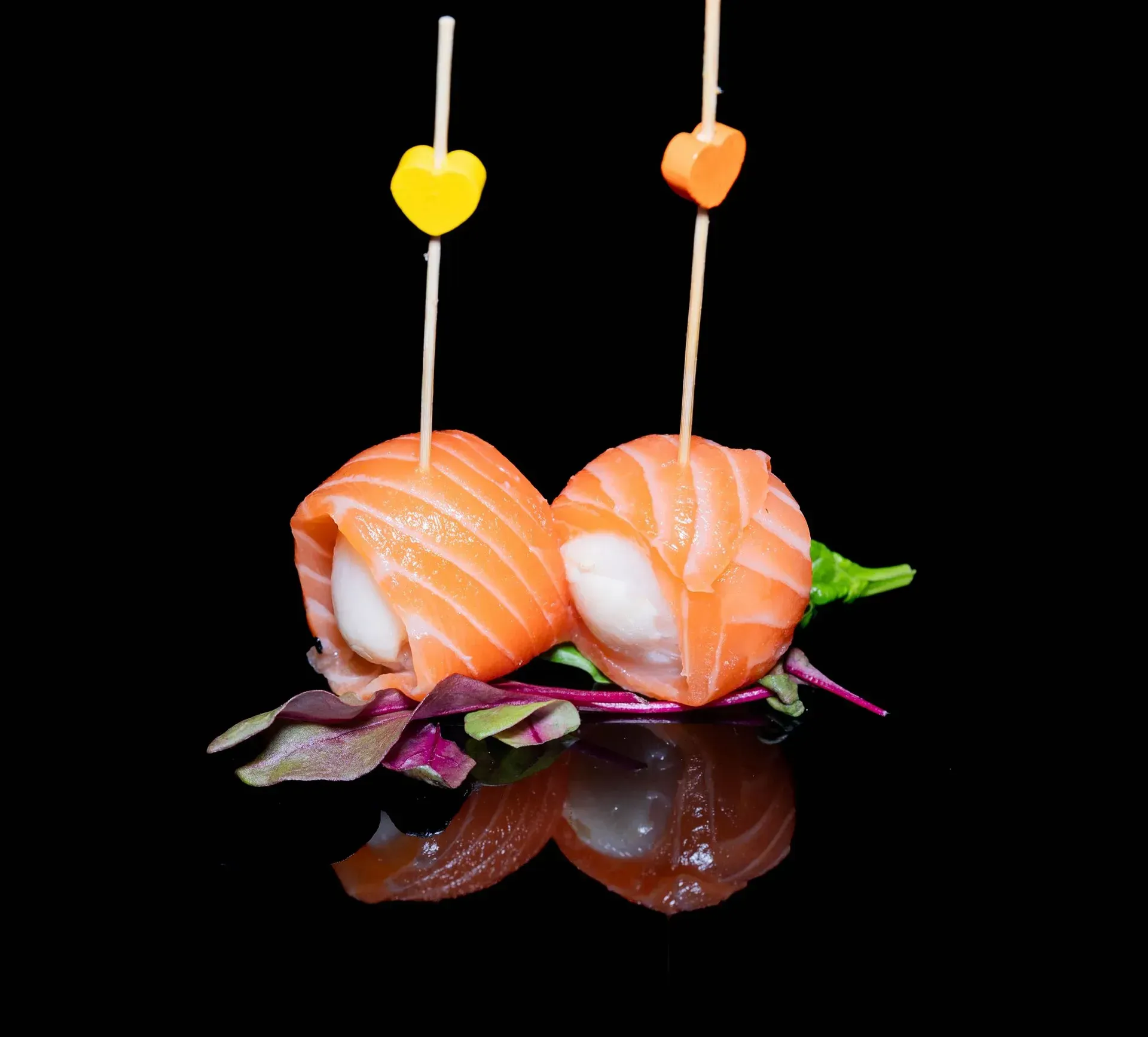 Due bocconcini di sushi di salmone su spiedini con formine a forma di cuore, serviti su una superficie nera riflettente.