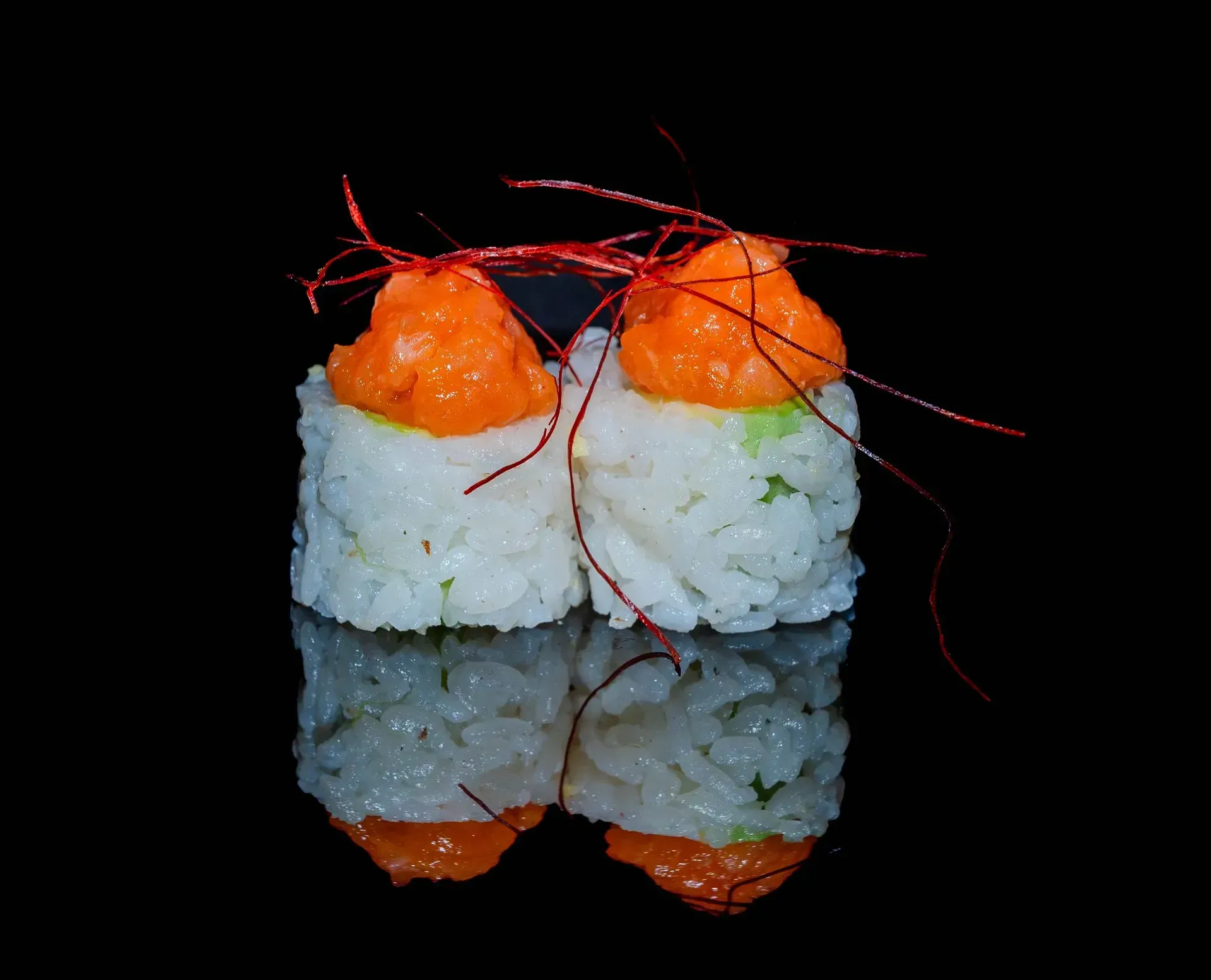 Due rotoli di sushi guarniti con uova di pesce arancioni e una sottile guarnizione rossa su una superficie nera riflettente.