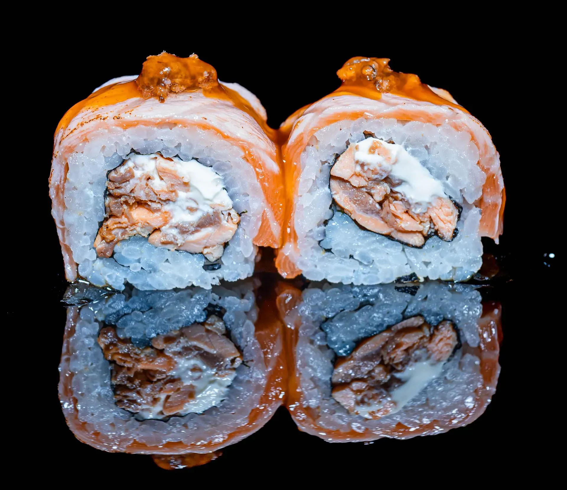 Due rotoli di sushi con salmone e ripieno cremoso, conditi con salsa all'arancia, su una superficie nera riflettente.