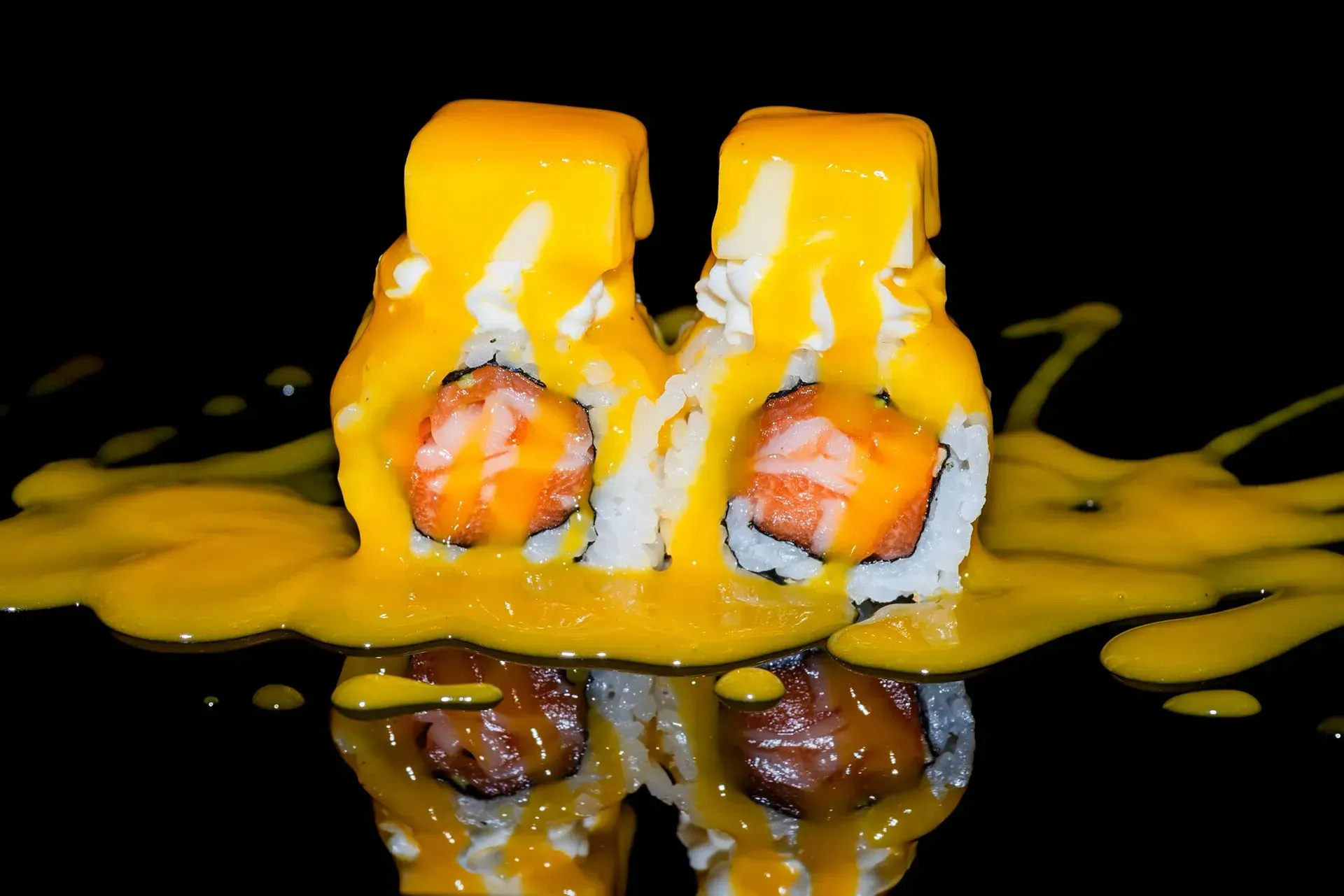 Due rotoli di sushi ricoperti di salsa all'arancia, che gocciola su una superficie nera con riflessi