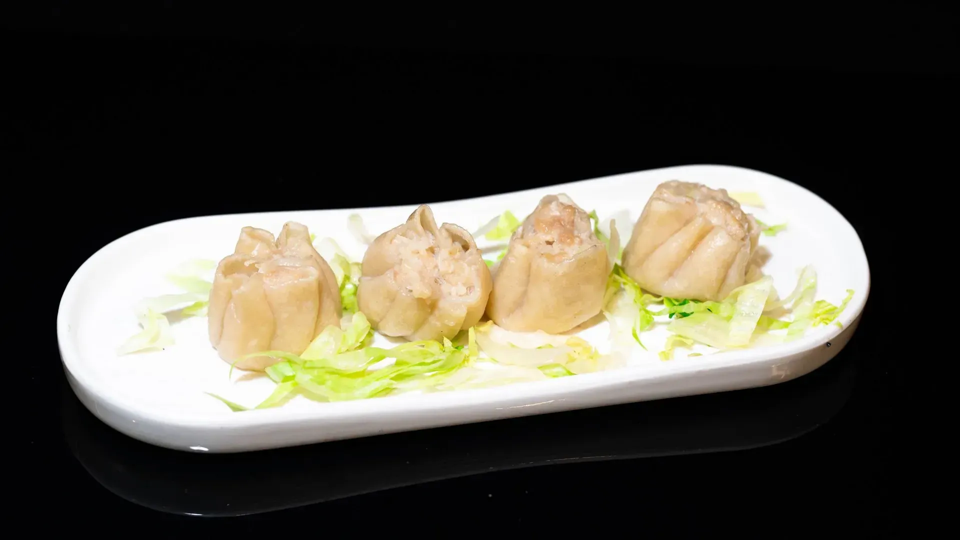 Quattro ravioli al vapore su un piatto bianco, guarniti con cipollotti verdi tritati su sfondo nero.